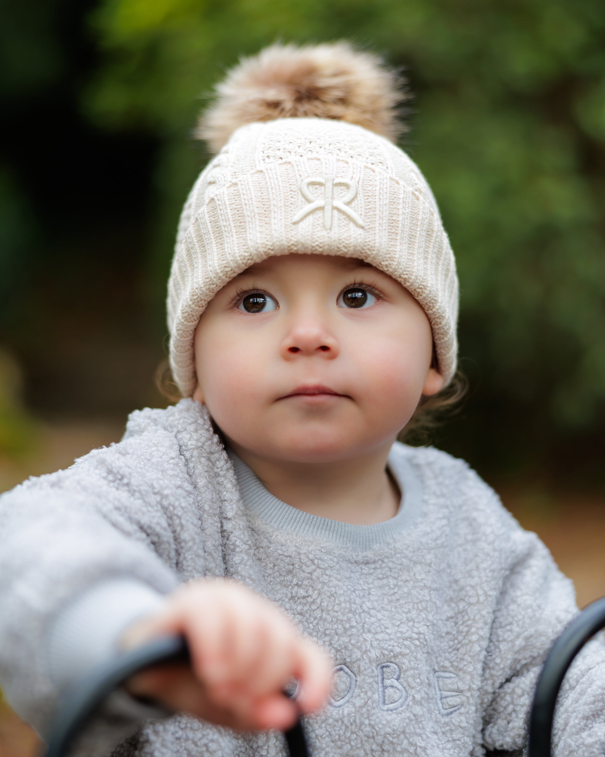 Child wearing a cream-colored knit bobble beanie hat with a pom-pom, and blue oversized teddy one-piece outdoors.