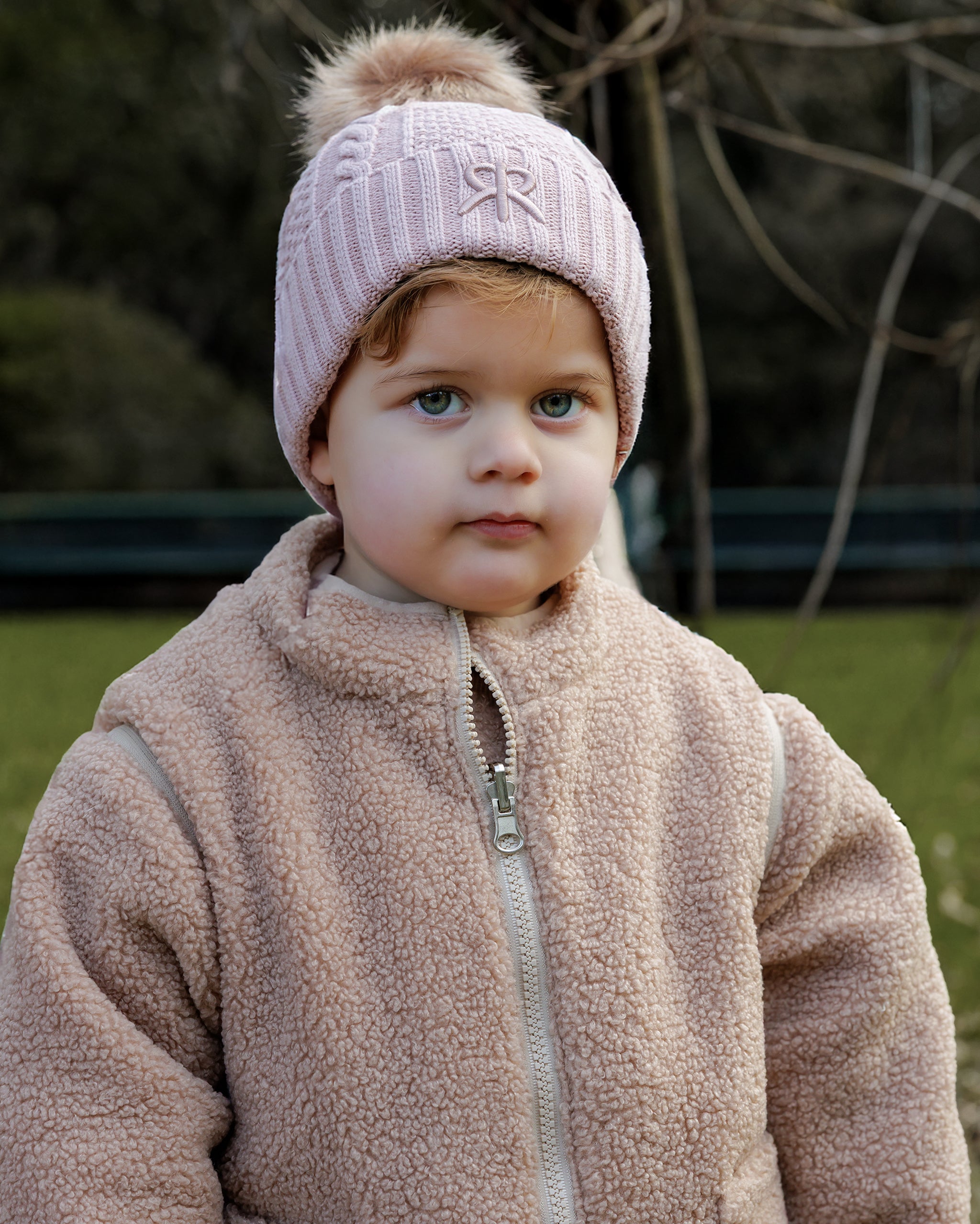 Child wearing a pink knit bobble beanie hat and 4-in-1 brown reversible teddy fleece jacket outdoors