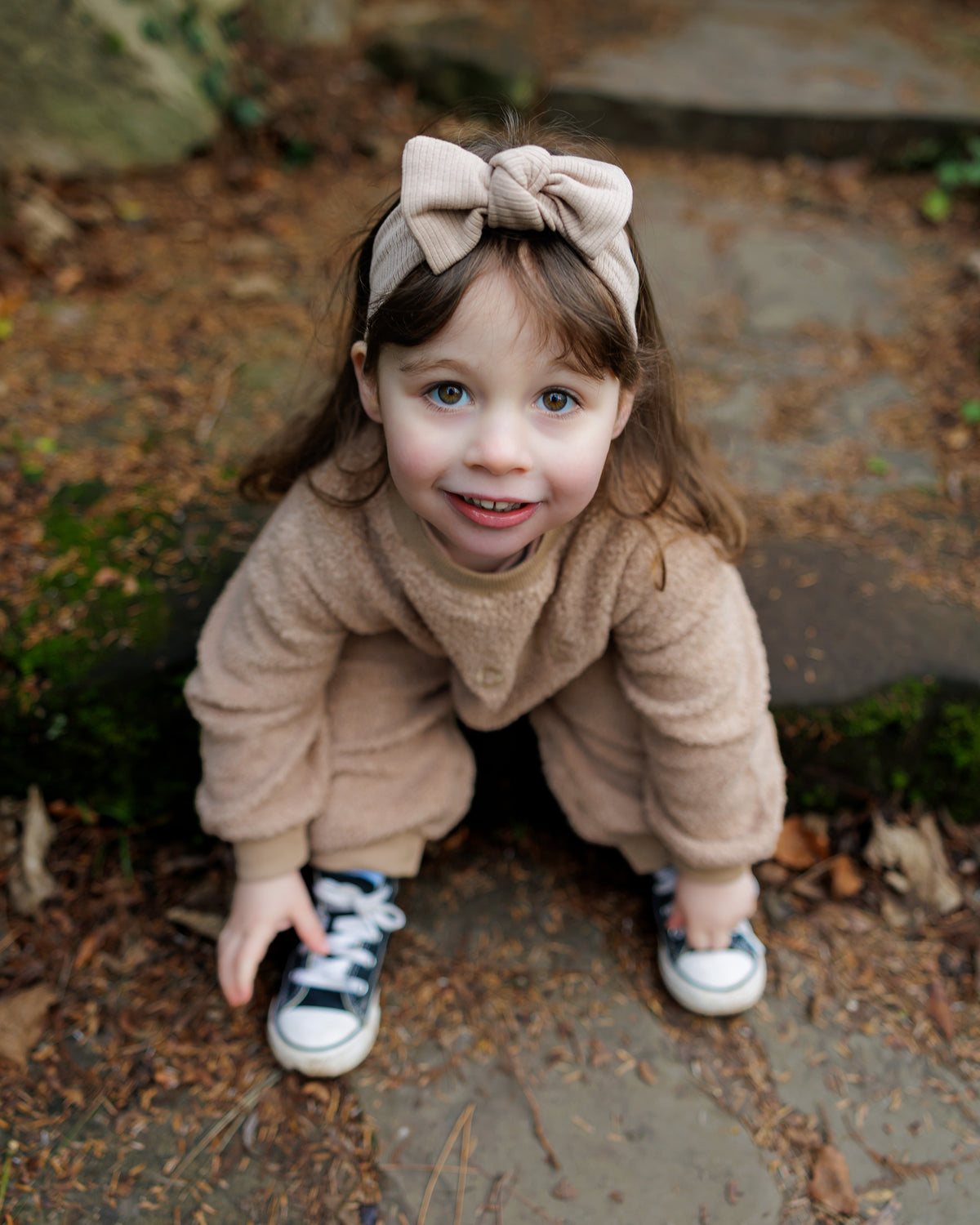 Child wearing a brown teddy one-piece outfit with a ribbed bow headband, standing outdoors on a stone path.