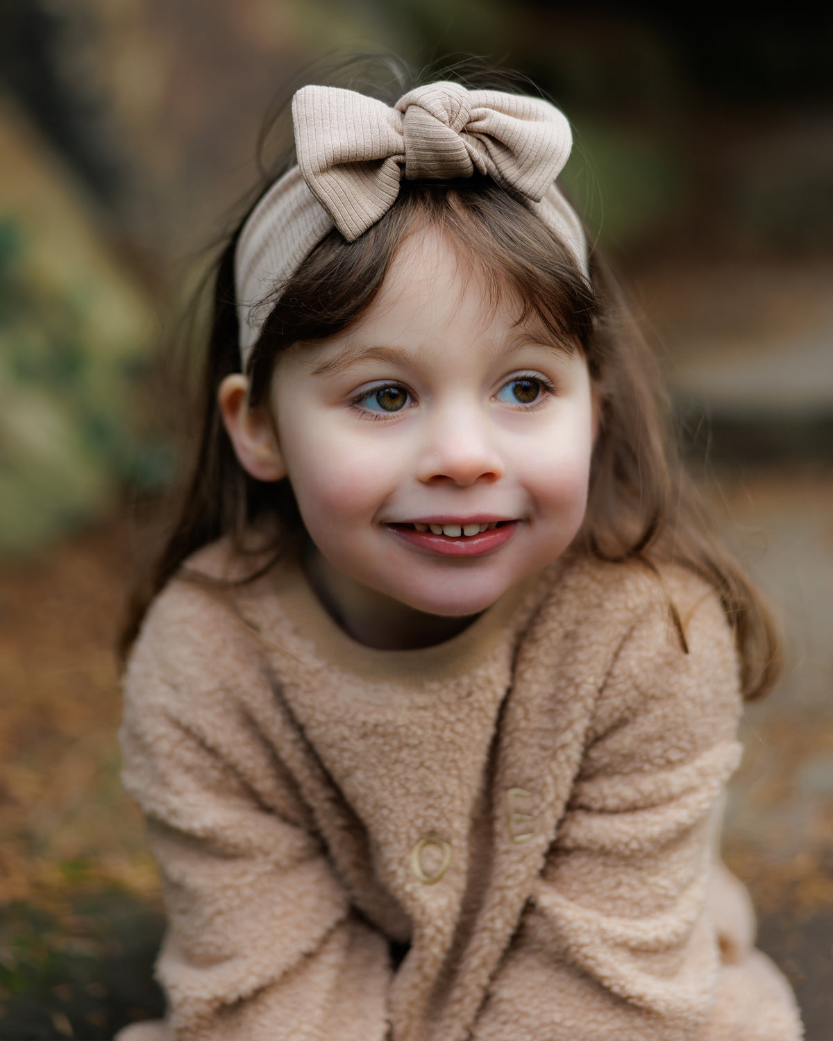 Child wearing a beige ribbed headband with a bow and teddy fleece outfit smiling outdoors.