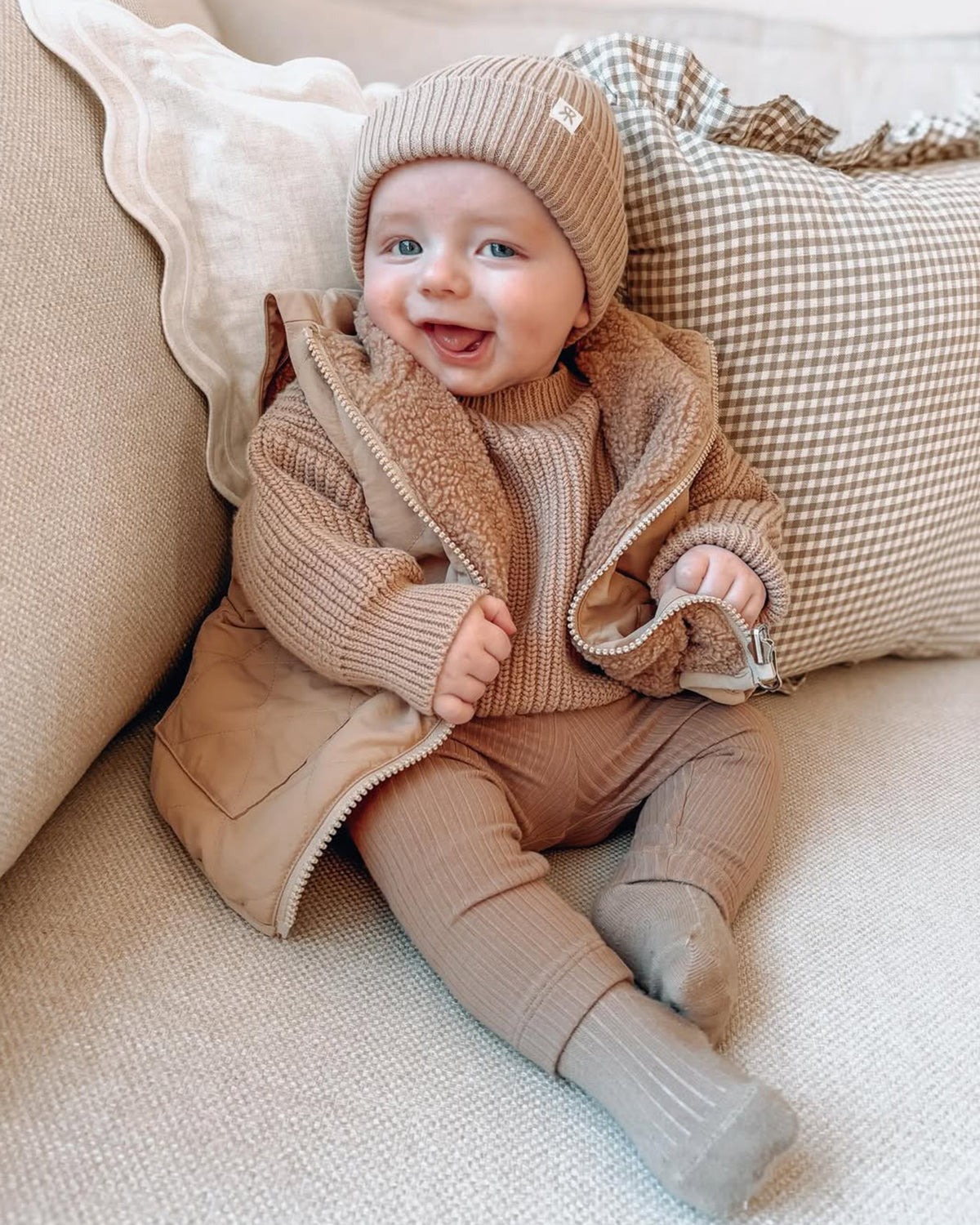 Baby in a brown 4-in-1 Reversible Coat/Gilet, chunky knit sweater jumper matching ribbed leggings, sitting on a couch with pillows