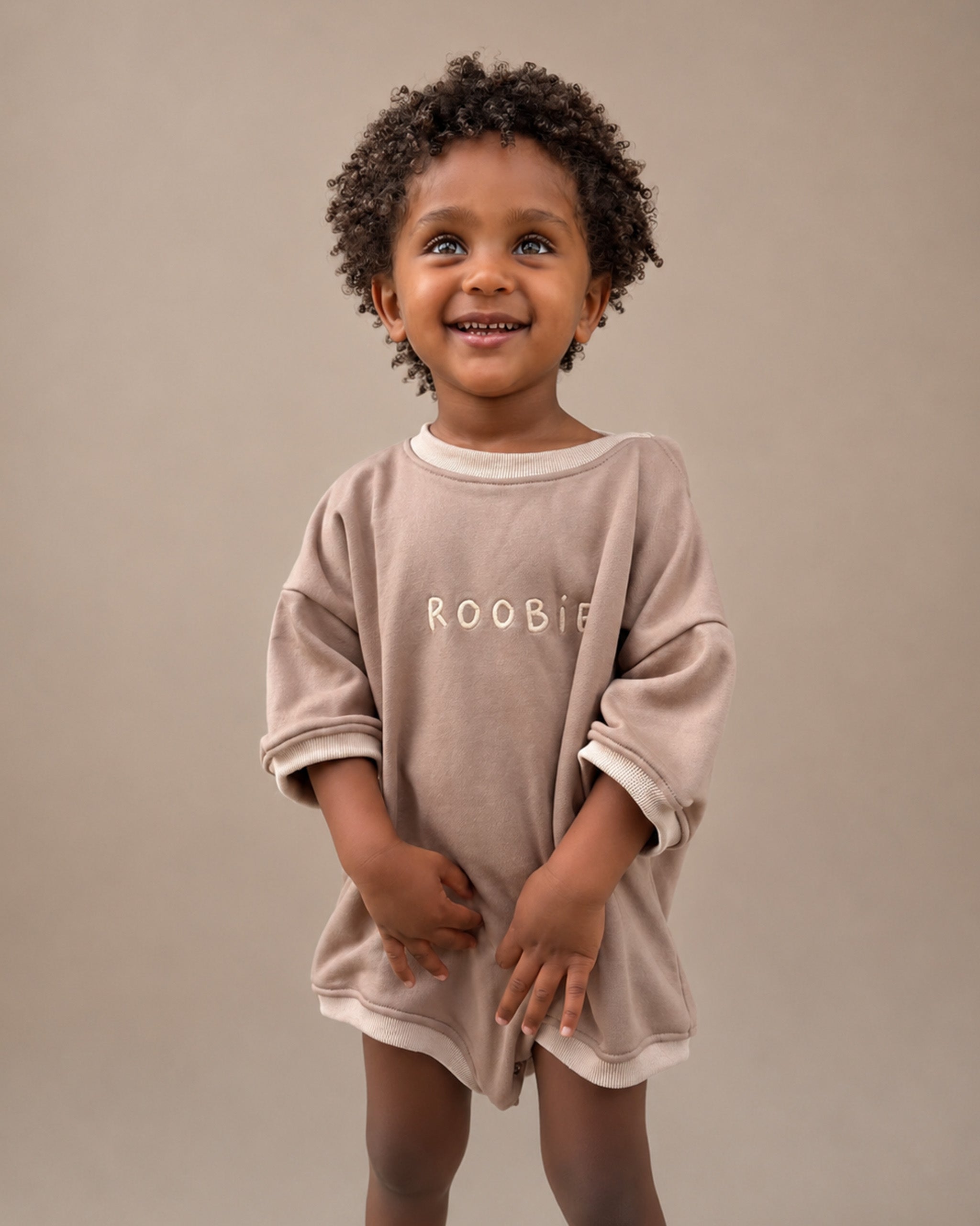 A smiling young child with curly dark hair stands against a plain warm beige studio background, wearing an oversized light taupe Romper with the name 'ROOBIE' printed across the chest.