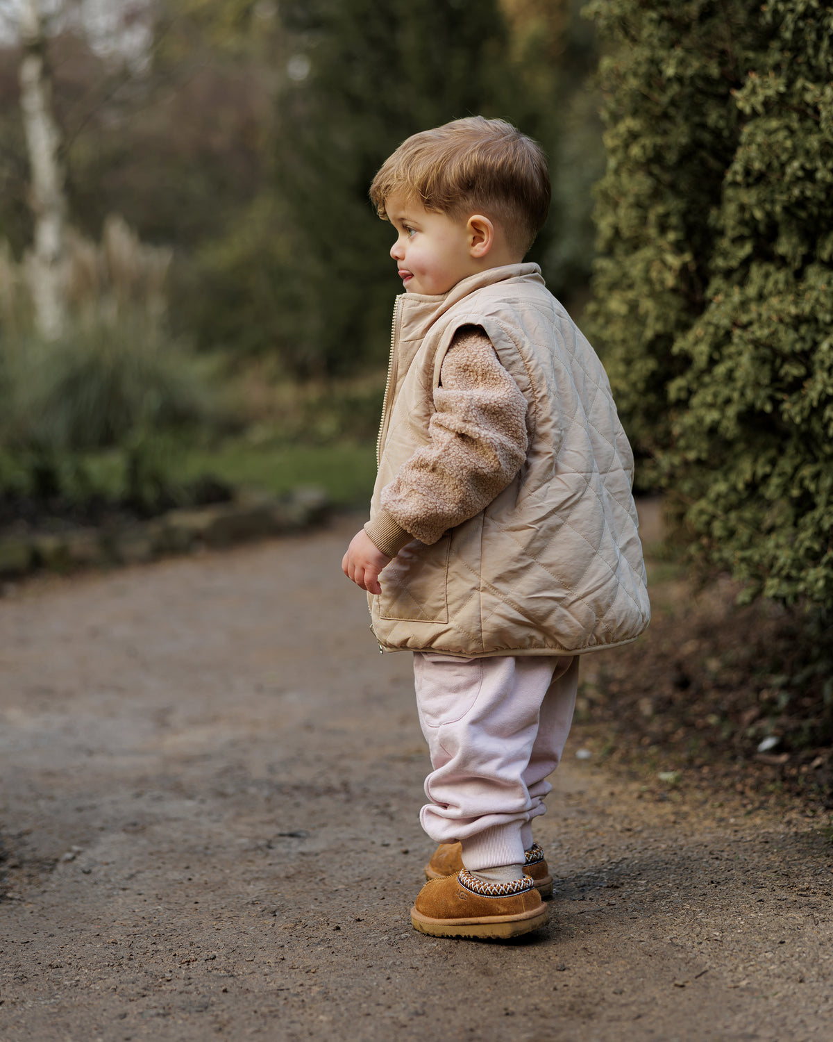 A toddler wearing soft pink ROOBiE cargo trousers showing the cargo pockets and oversized fit with a quilted gilet and brown teddy jumper, outdoors.