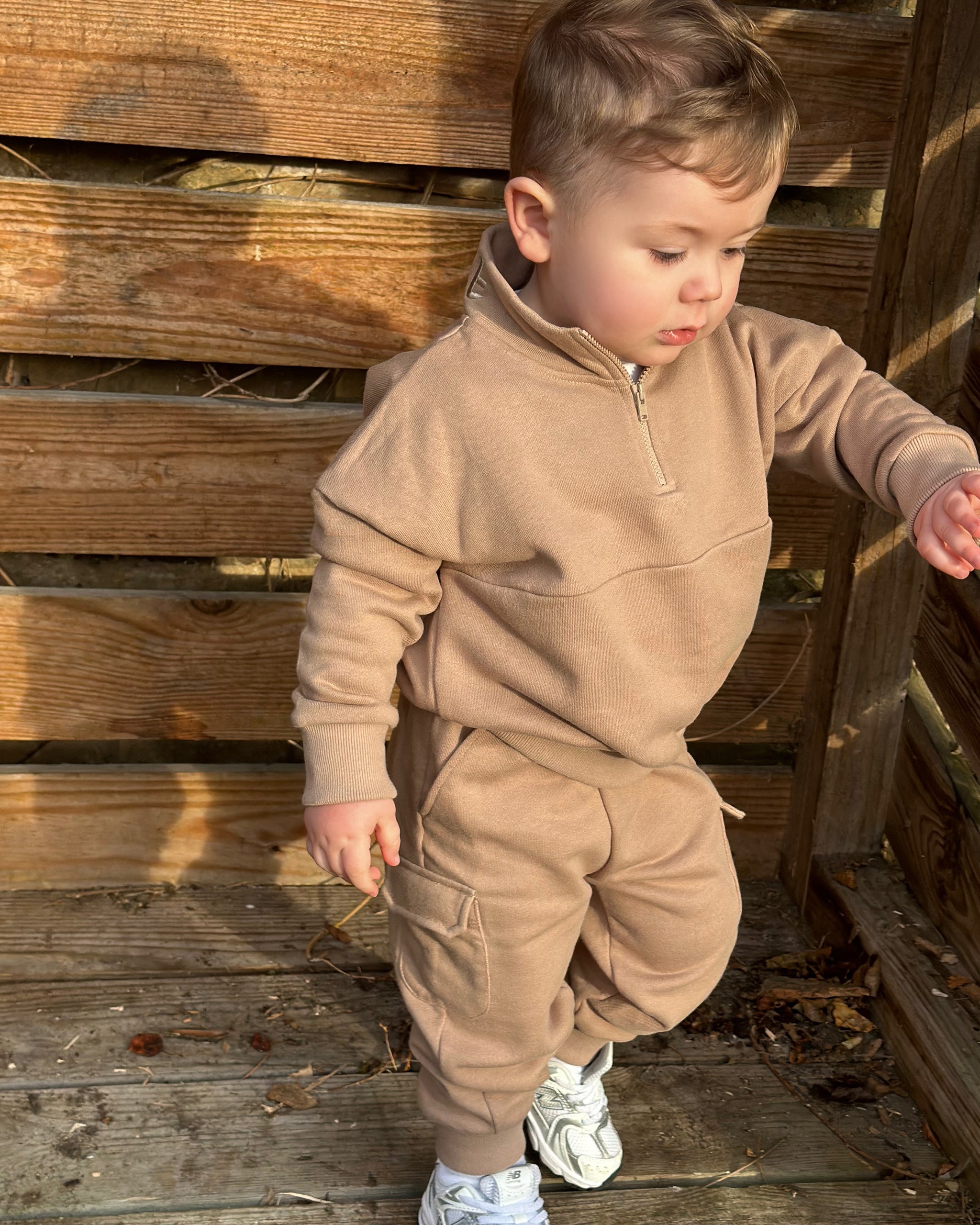 Child in a brown tracksuit with quarter zip and matching cargo pants standing against a wooden wall