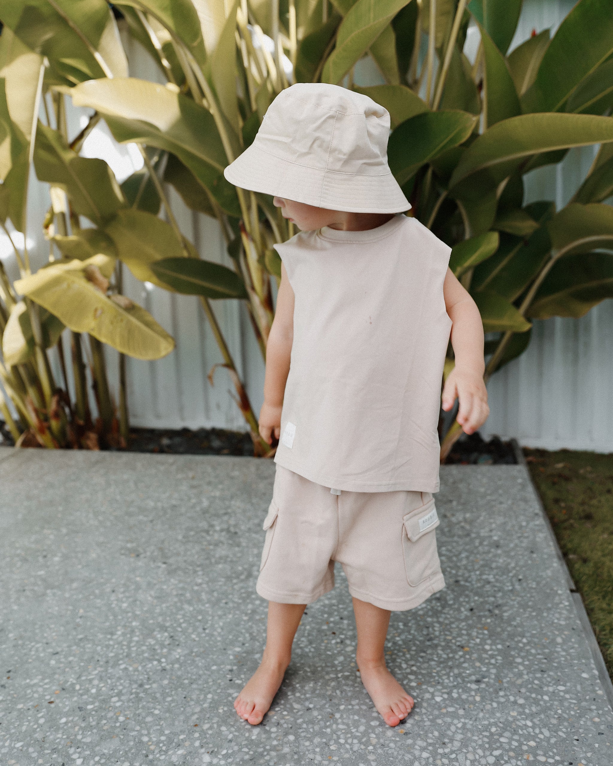 A toddler standing barefoot outdoors on a paved surface, wearing a light beige sleeveless Singlet top, matching cargo shorts, and a cream neutral reversible bucket hat in front of large green tropical leaves.