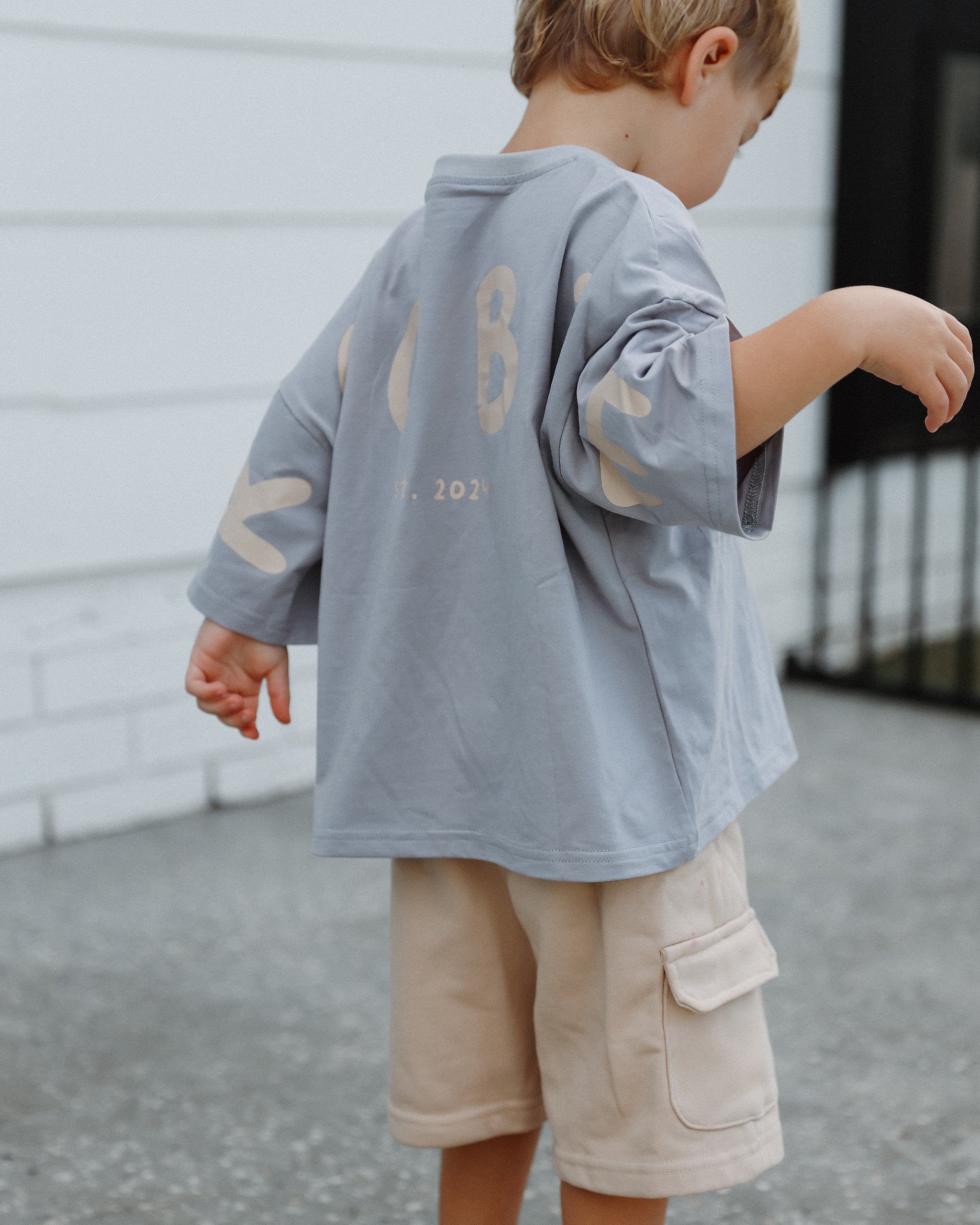 A young child mixing and matching a light blue Oversized T-shirt and cream Cargo Shorts, standing outdoors in front of a neutral white building with beige graphic lettering spelling "ROOBiE" on the back