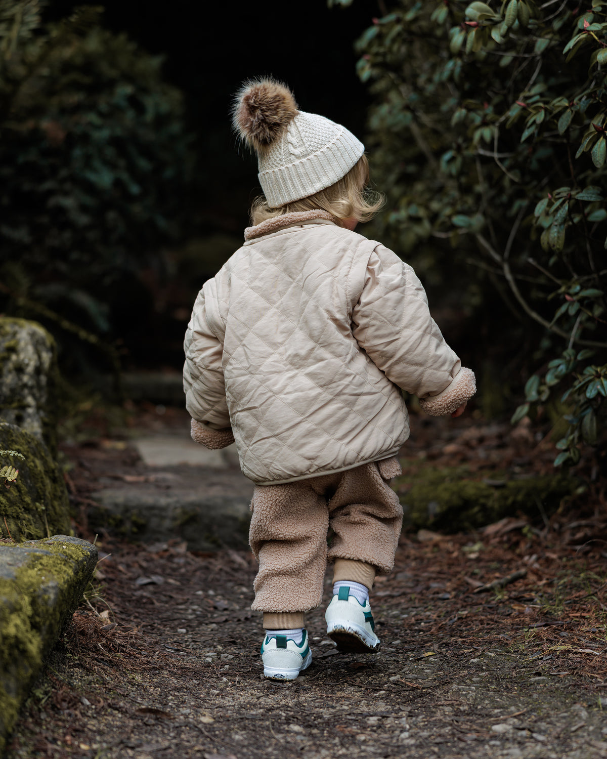 Child in a beige quilted reversible coat jacket and cream knit bobble beanie hat and brown teddy cargo trousers walking on a path surrounded by greenery