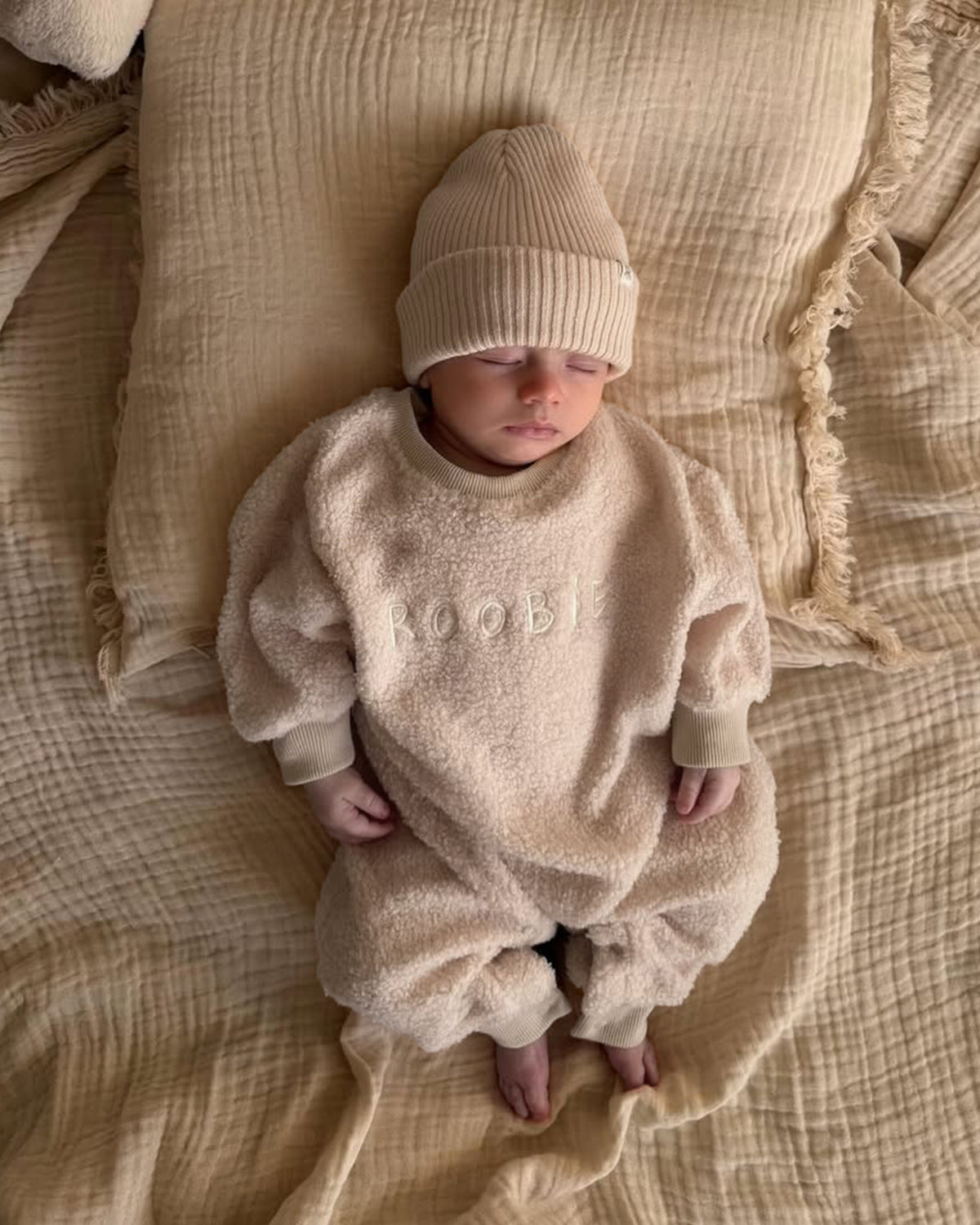 Newborn baby in a cream teddy one-piece and matching knit cotton beanie lying on a neutral bedding.