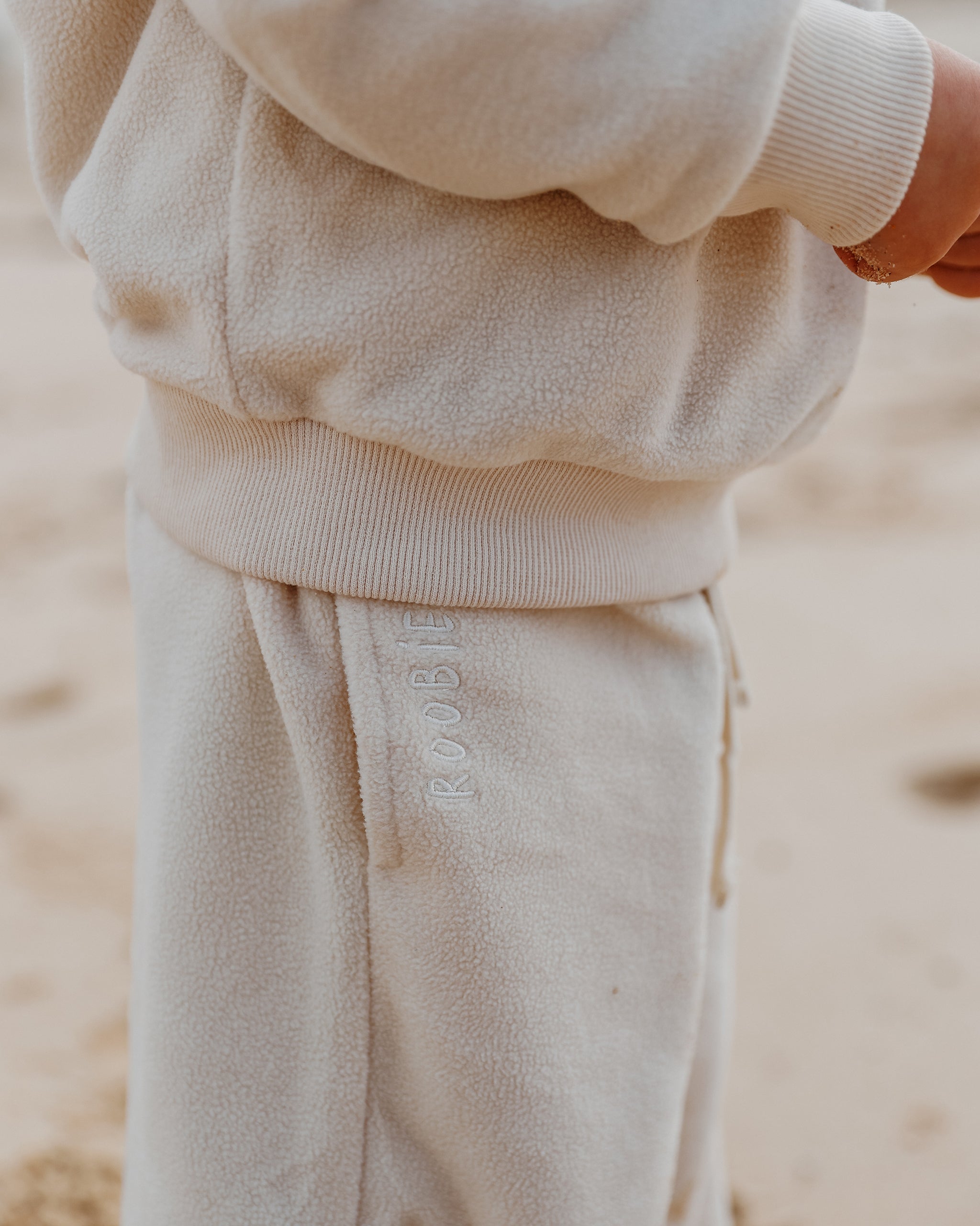 A close-up of a child wearing a cream Fleece Quarter Zip Jumper and matching Fleece Wide Leg Joggers with 'ROOBiE' embroidered vertically on the thigh, set against a warm neutral, sandy background.