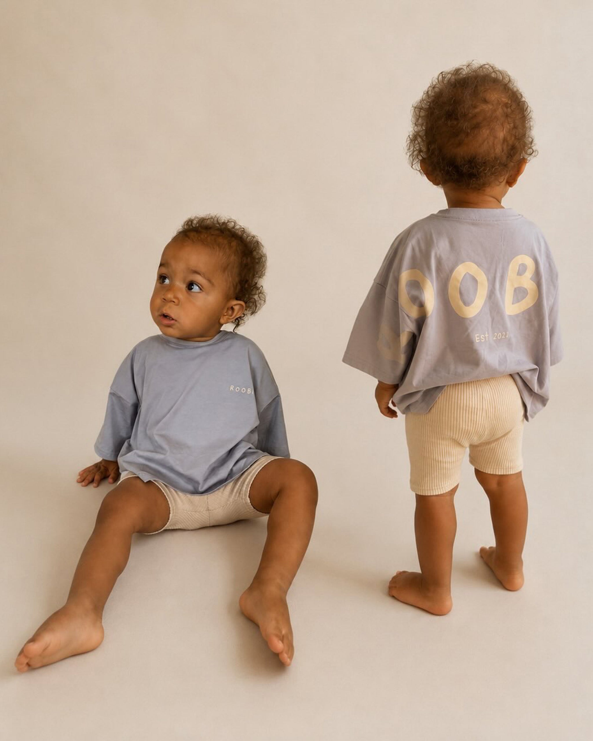 Two toddlers modeling matching light blue Oversized T-Shirts and cream Ribbed Cycling Shorts against a warm beige studio backdrop. One toddler is seated facing left, and the other stands with their back to the camera, showing a large cream 'ROOBiE' logo on the back of the shirt.