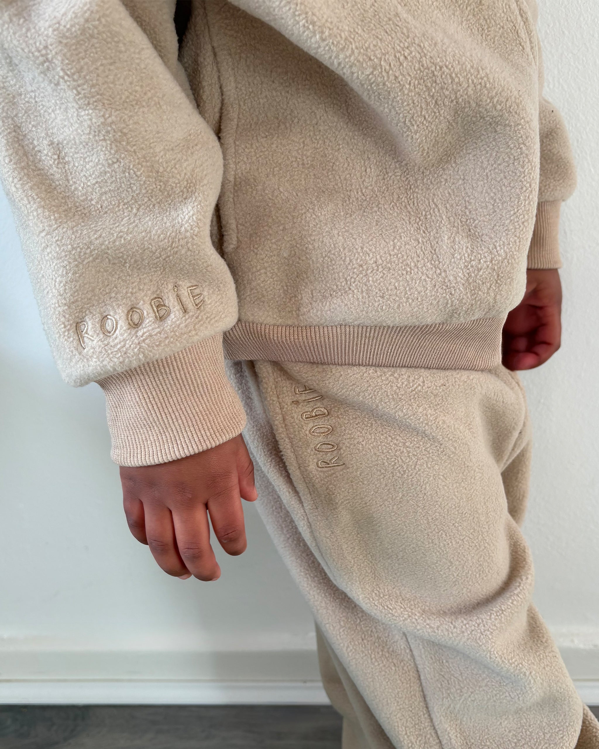 Beige fleece tracksuit worn by a toddler with embroidery wrist and leg details showing brand logo against a neutral background