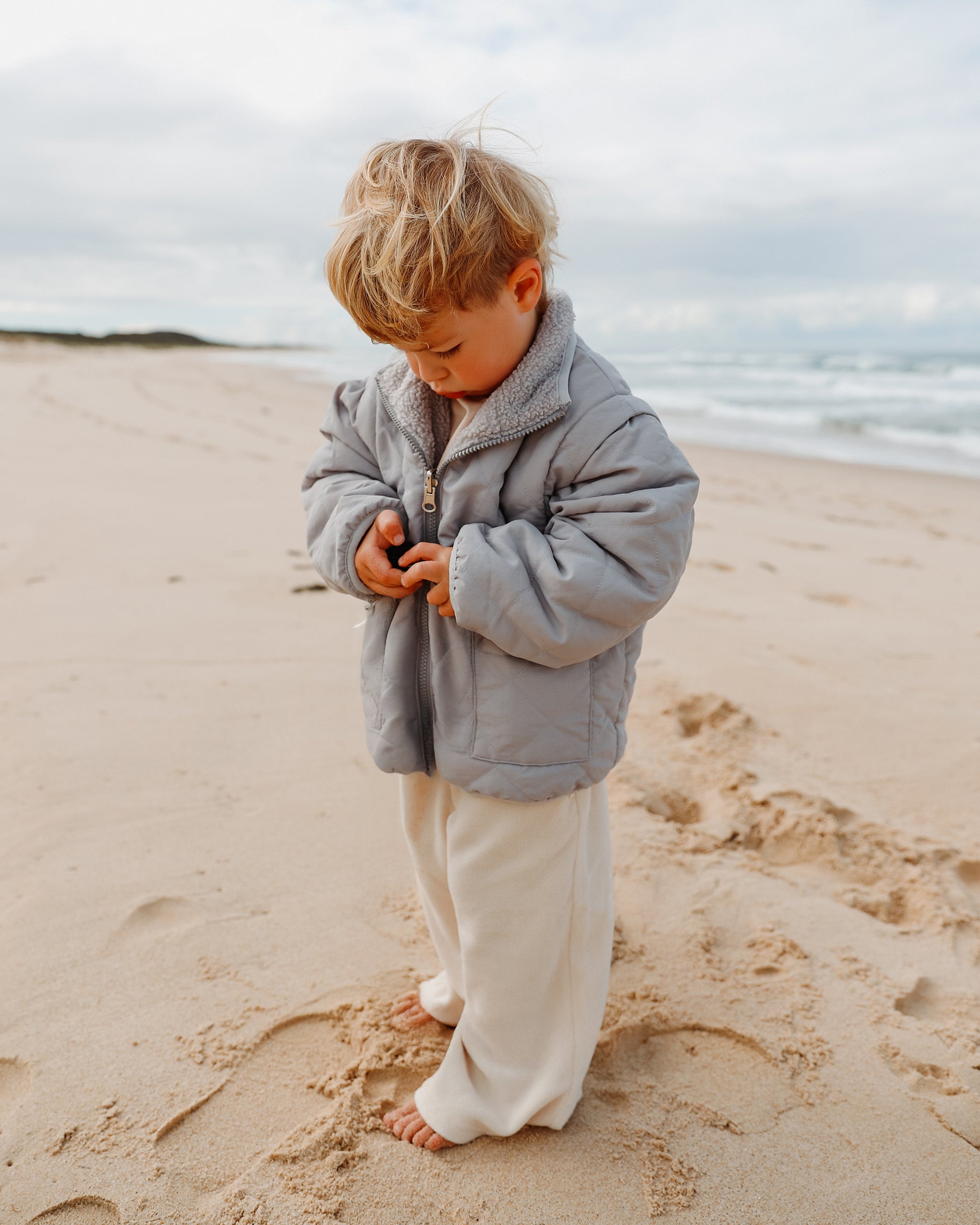 A young child standing on a sandy beach wearing a light gray-blue 4-in-1 reversible teddy jacket and cream-colored wide leg jogger pants, looking down while buttoning the jacket.