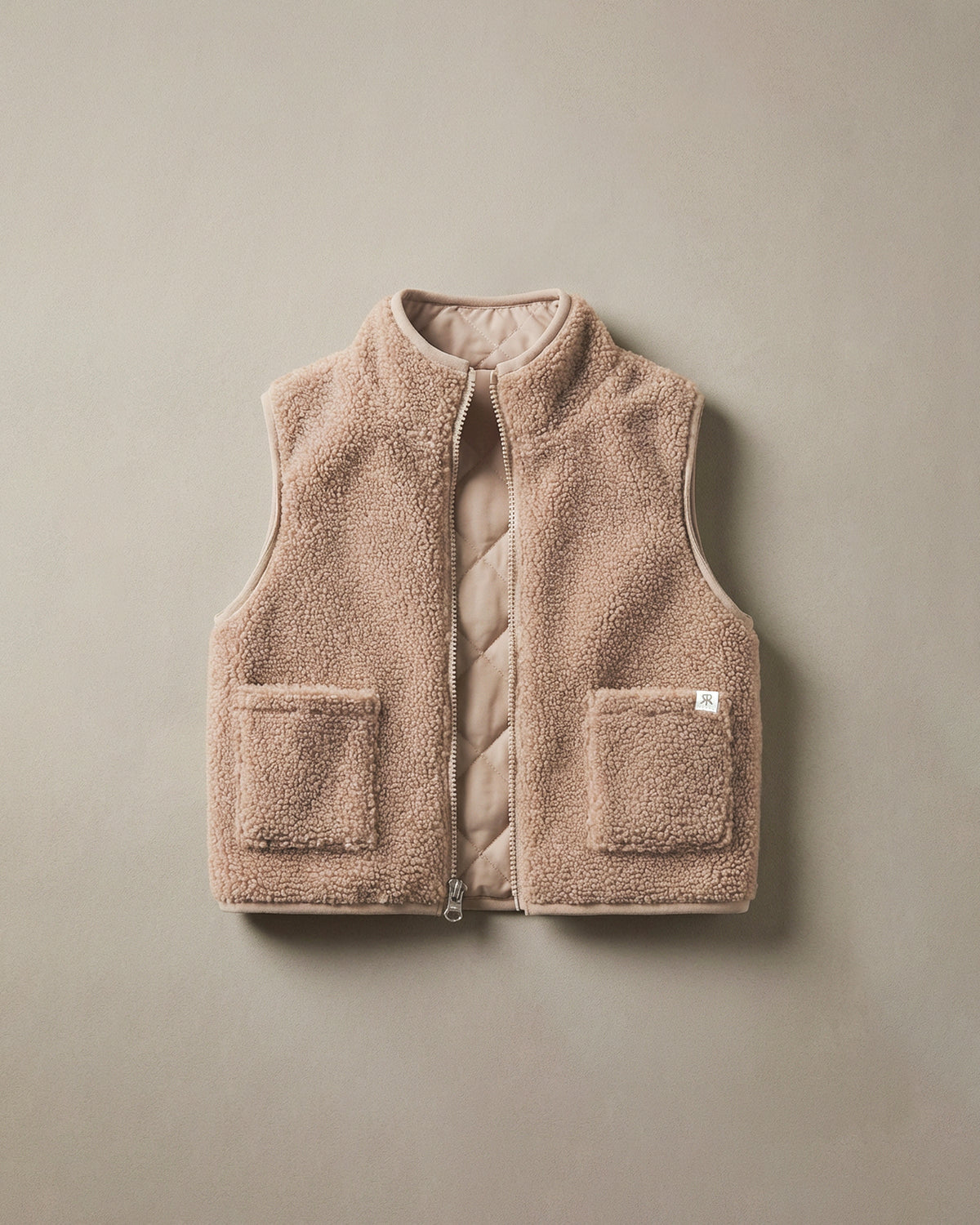 A sleeveless teddy fleece gilet in a warm taupe-beige color, featuring a quilted interior lining, a front zipper, and two front patch pockets, shown flat against a neutral beige background.