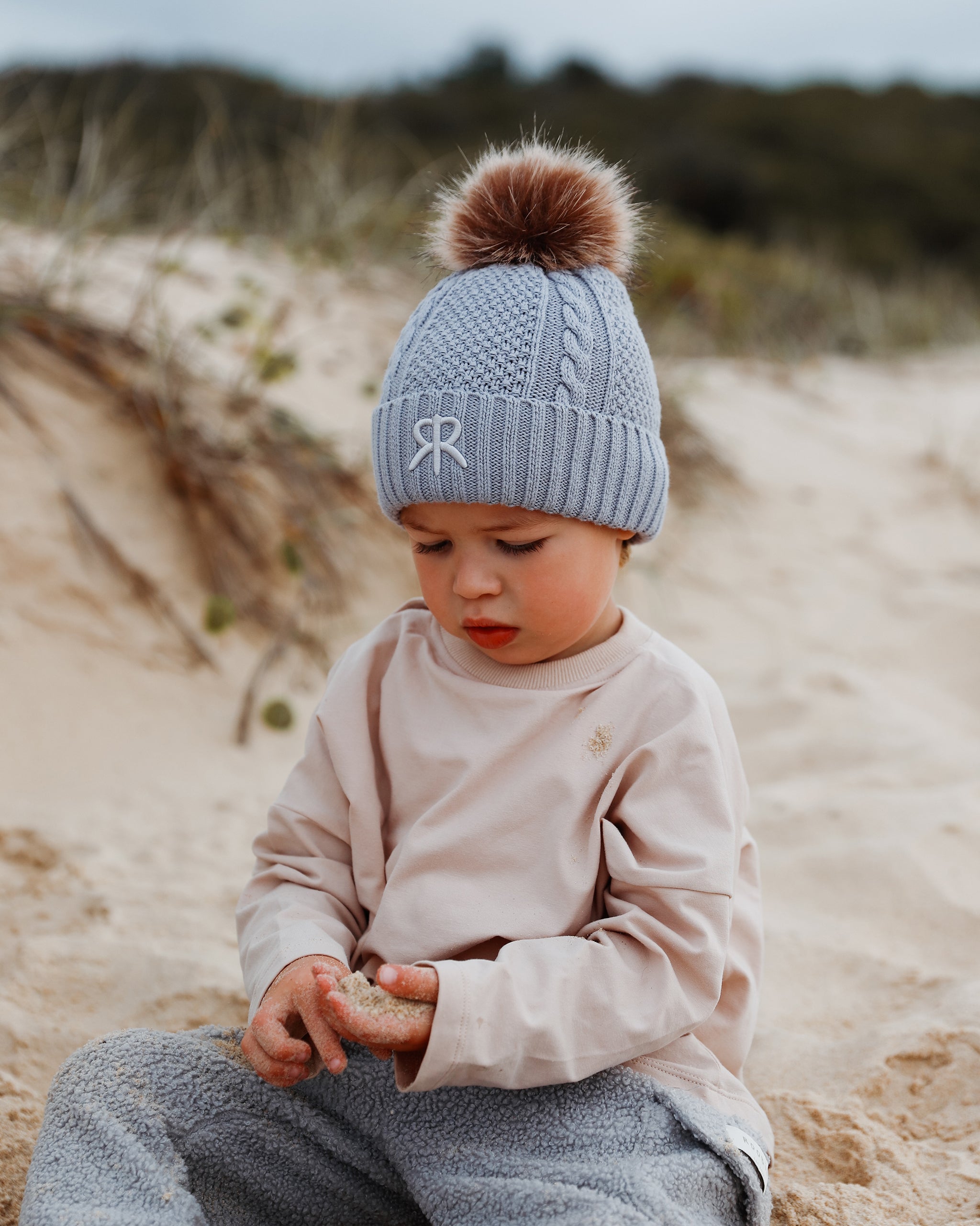 A toddler sitting on a sandy beach wearing a light blue cable-knit bobble beanie with a brown pom-pom and a neutral embroidered monogram logo, paired with a beige long-sleeve top and gray-blue teddy cargo pants.
