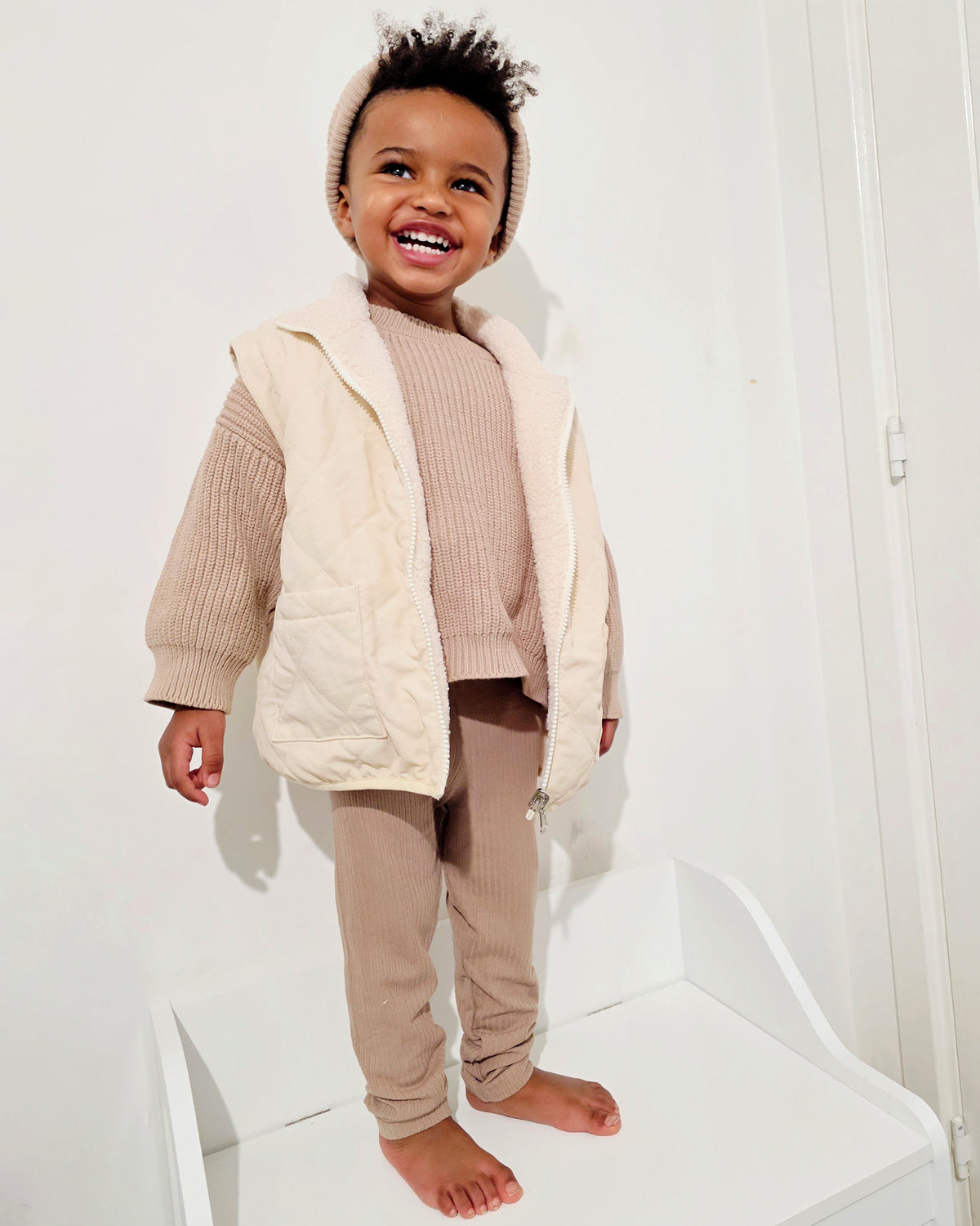 Child wearing a brown chunky knit sweater, 4-in-1 reversible quilted teddy coat/gilet, and matching ribbed leggings against a white background