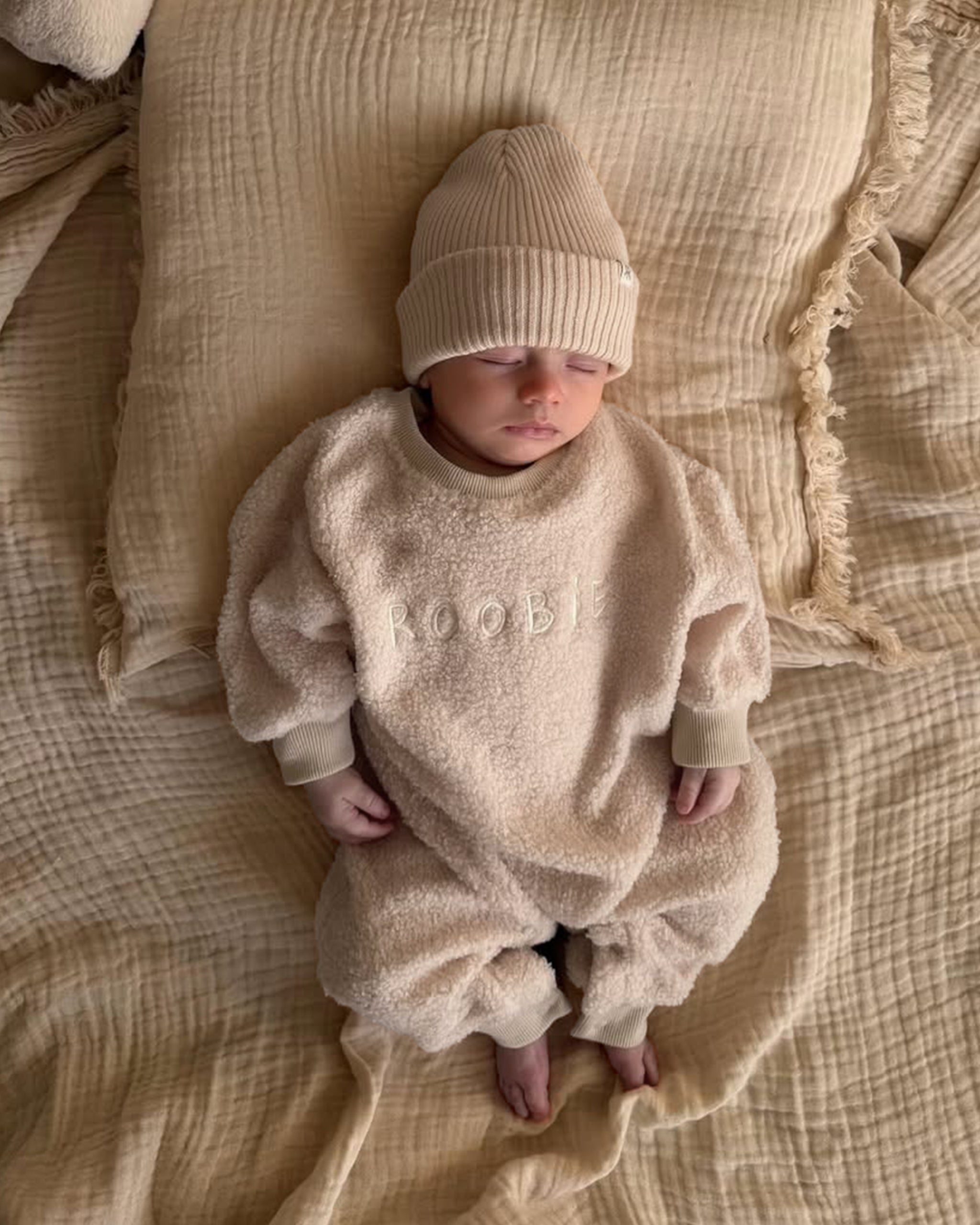 Newborn baby in a beige teddy onesie style one-piece and matching ribbed cotton beanie hat lying on neutral bedding.