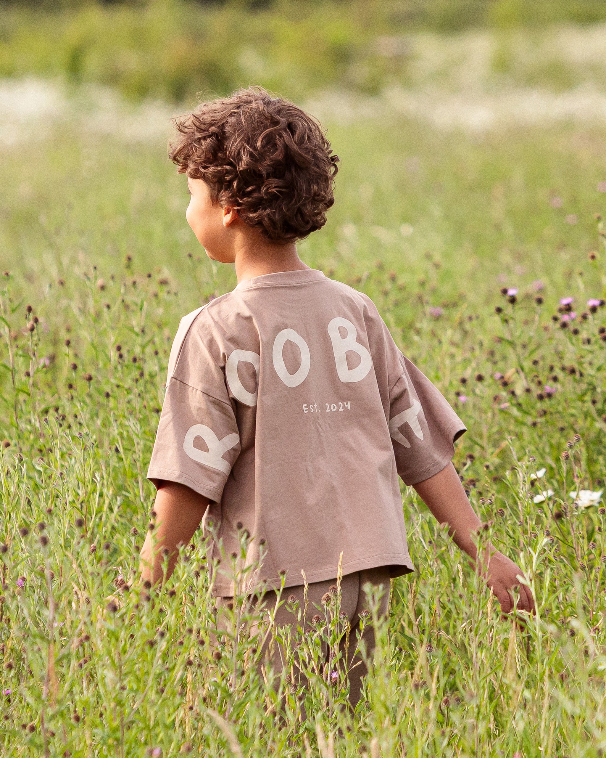 A young child with curly brown hair stands in a grassy field, viewed from behind, wearing a taupe-brown Oversized T-shirt with cream graphic lettering saying 'ROOBiE' on the back.