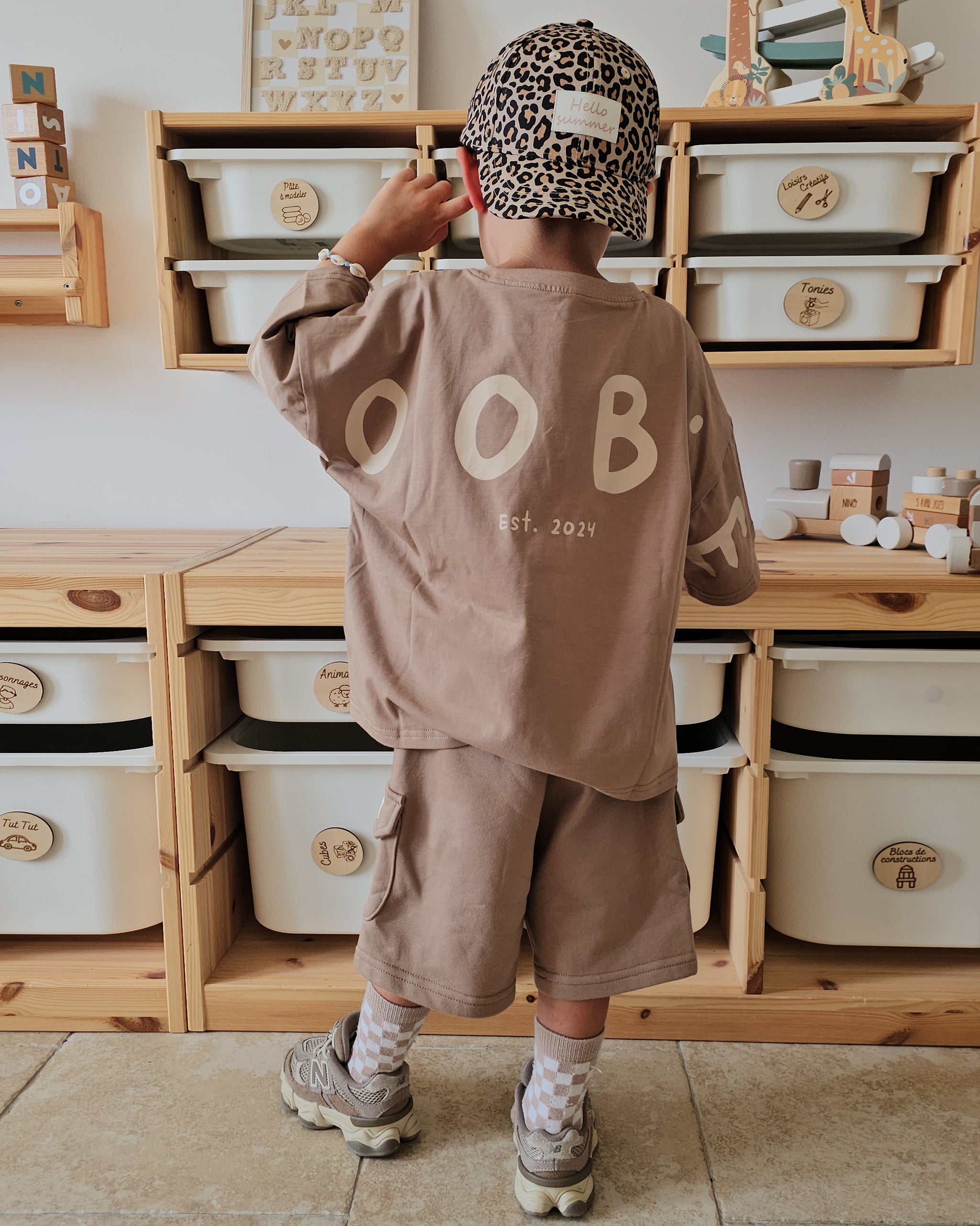 A child standing with their back to the camera, wearing a matching taupe-brown Oversized T-Shirt and Cargo Shorts co-ord set with a leopard-print cap. The shirt features the letters 'ROOBiE' in a cream colour on the back.