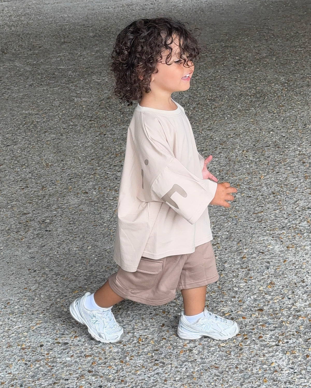 A toddler with curly dark hair is shown from the side walking outdoors on a gray asphalt surface. The child is wearing a light beige oversized t-shirt, brown cargo shorts, and white sneakers.