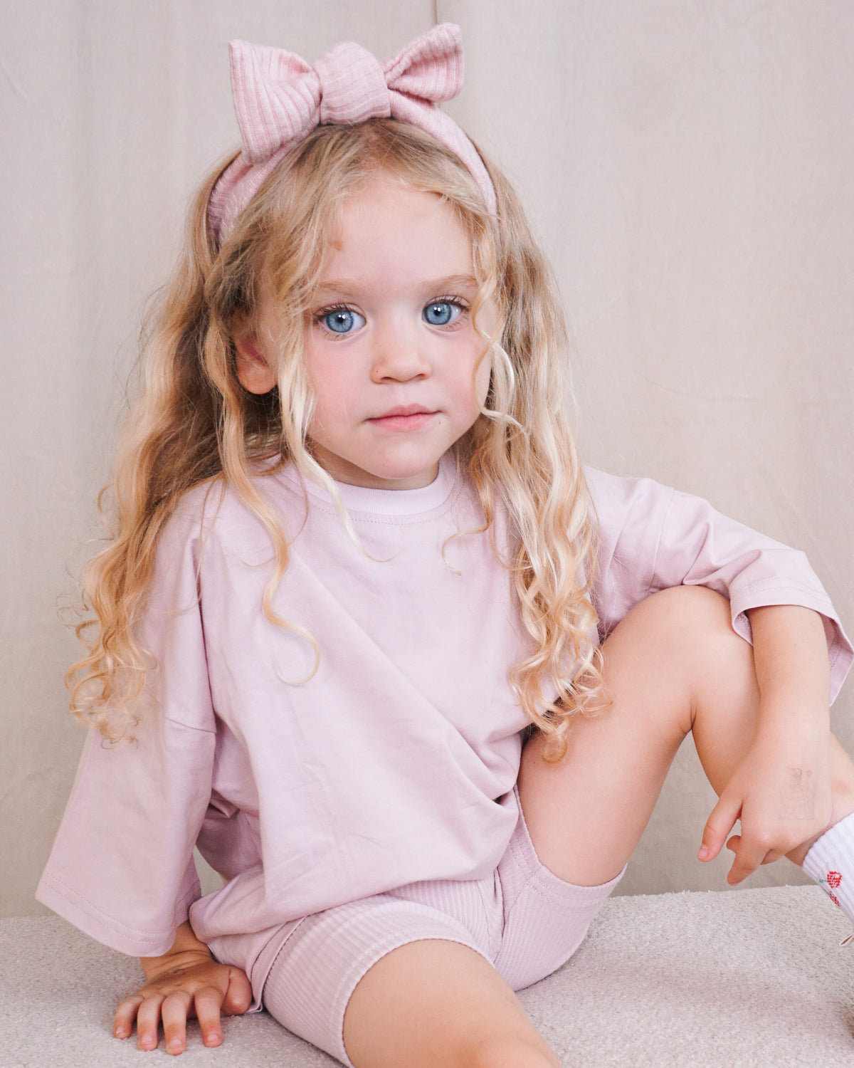 A young girl with long blonde curly hair sits on a light beige carpet wearing a matching light pink Oversized T-Shirt and Ribbed Cycling Shorts co-ord, along with a pink striped Ribbed Bow Headband.