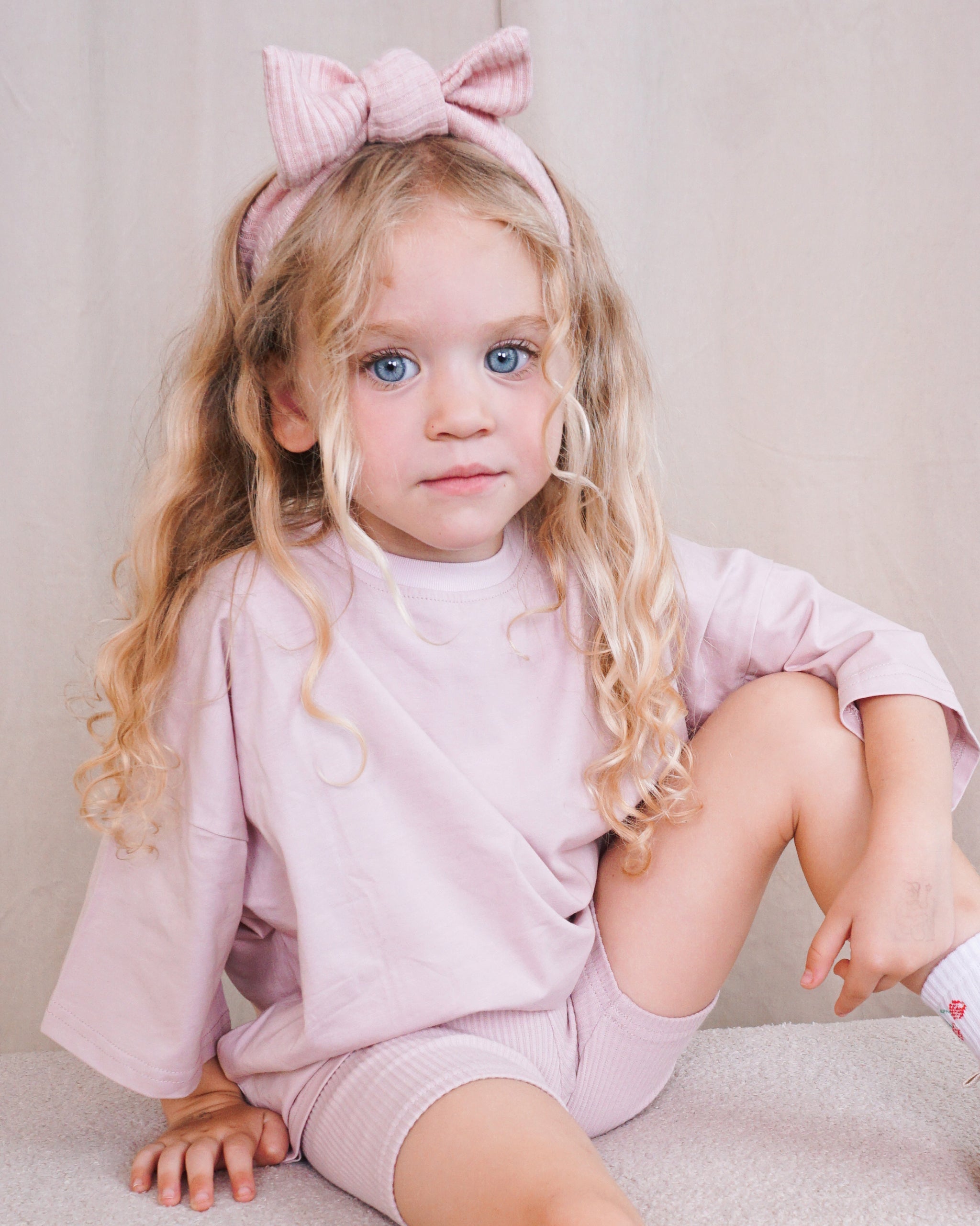 A young girl with long blonde curly hair sits on a light beige carpet wearing a matching light pink Oversized T-Shirt and Ribbed Cycling Shorts co-ord, along with a pink striped Ribbed Bow Headband.