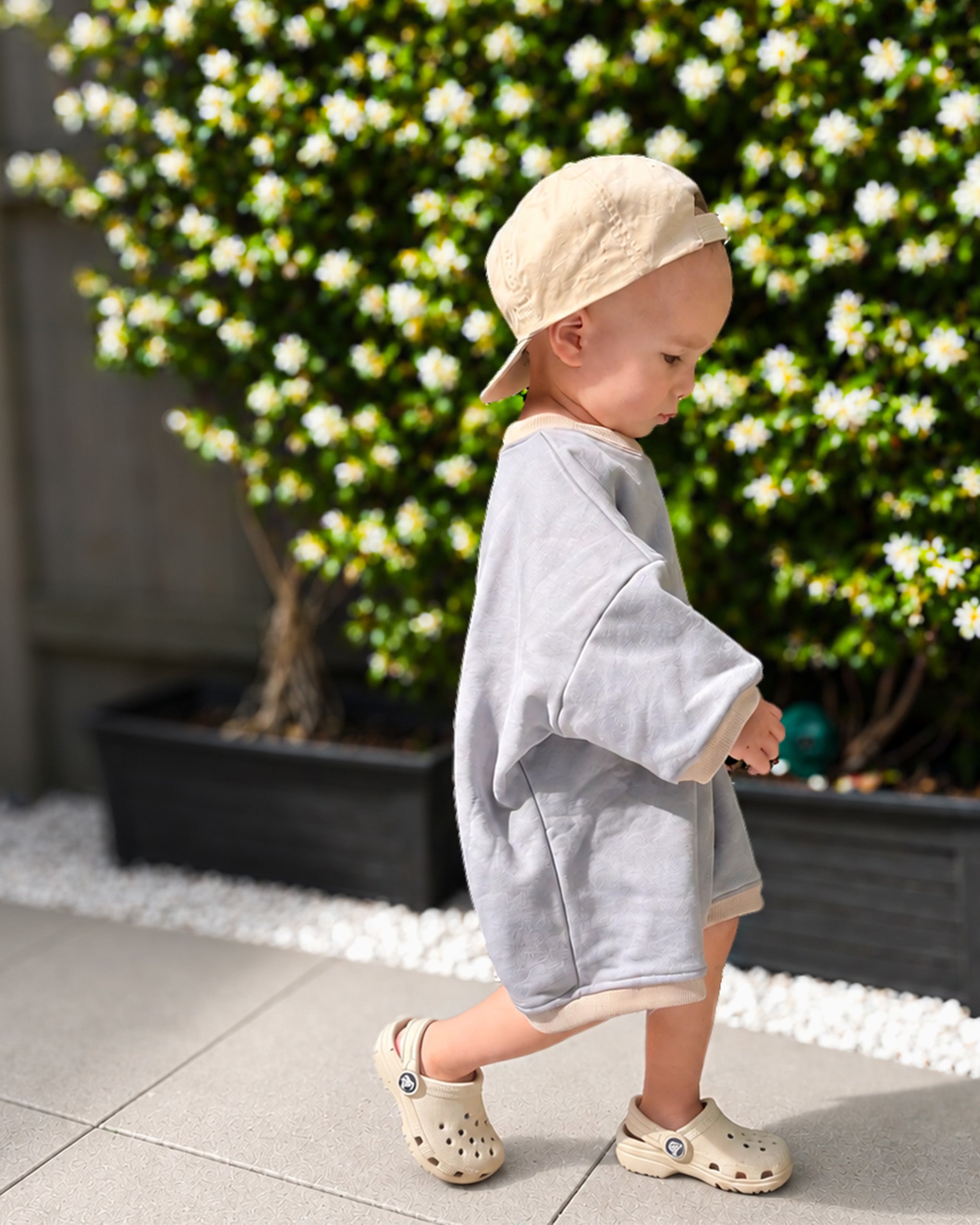 A toddler walking outdoors in a light gray-blue Oversized Romper, a beige Reversible Baseball Cap, and beige crocs, set against a background of green bushes with white flowers.