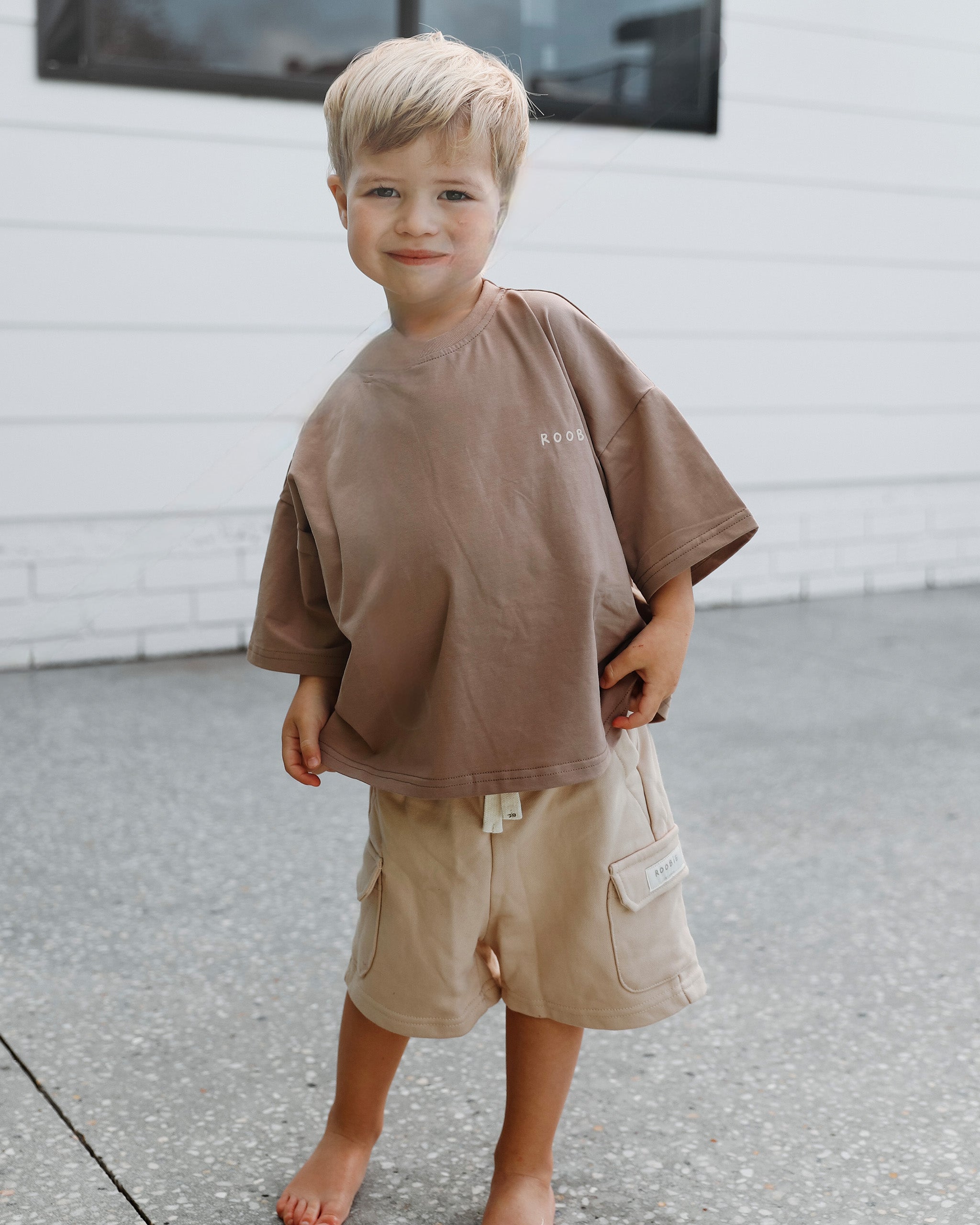 A young boy wearing an oversized taupe-brown t-shirt and beige-cream cargo shorts, standing barefoot on a concrete surface in front of a white brick wall.
