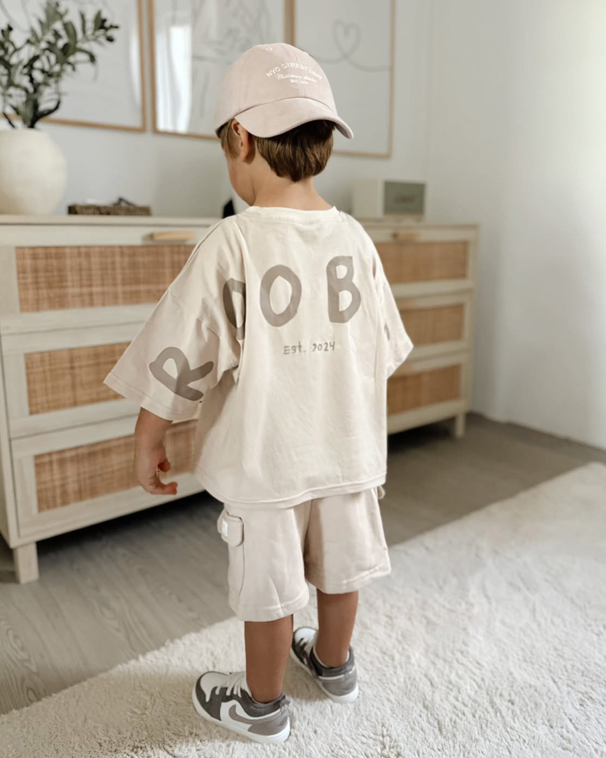 A toddler standing in a bright room, viewed from behind, wearing a cream-colored oversized t-shirt with a large brown 'ROOBiE' graphic on the back, matching cargo shorts, a neutral baseball cap, and gray sneakers.