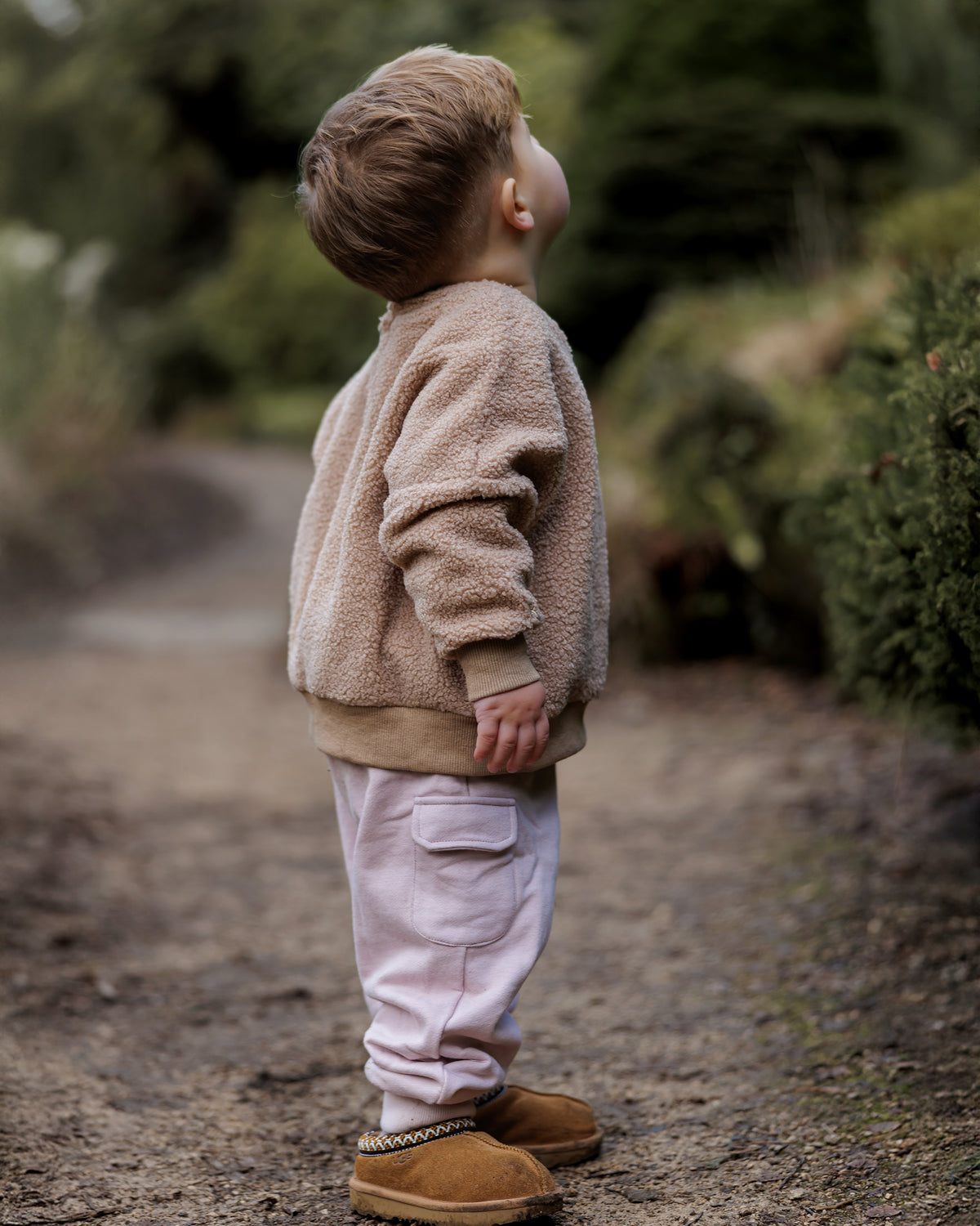 Child in a beige teddy fleece jumper and pink cargo pants standing on a forest path.