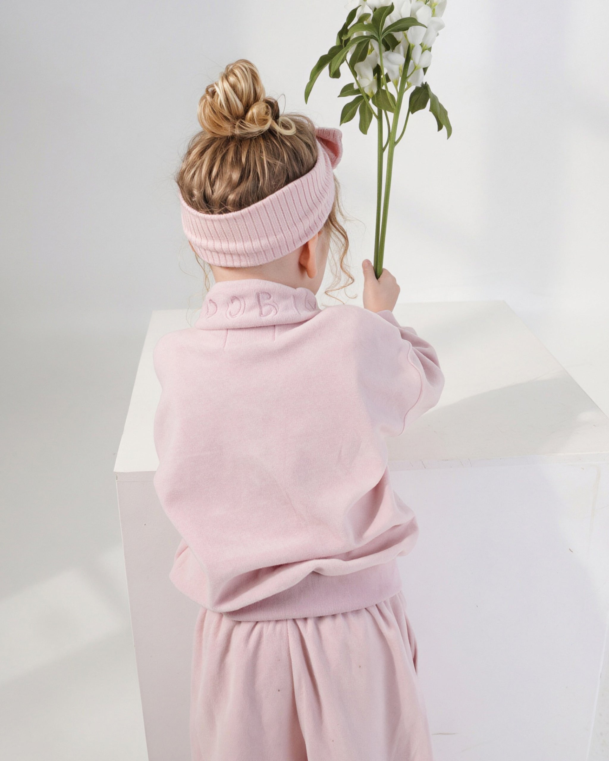 A child seen from behind wearing a light pink ribbed bow headband and a matching pale pink quarter zip jumper and wide leg jogger set, holding a green stem with white flowers against a white studio background.