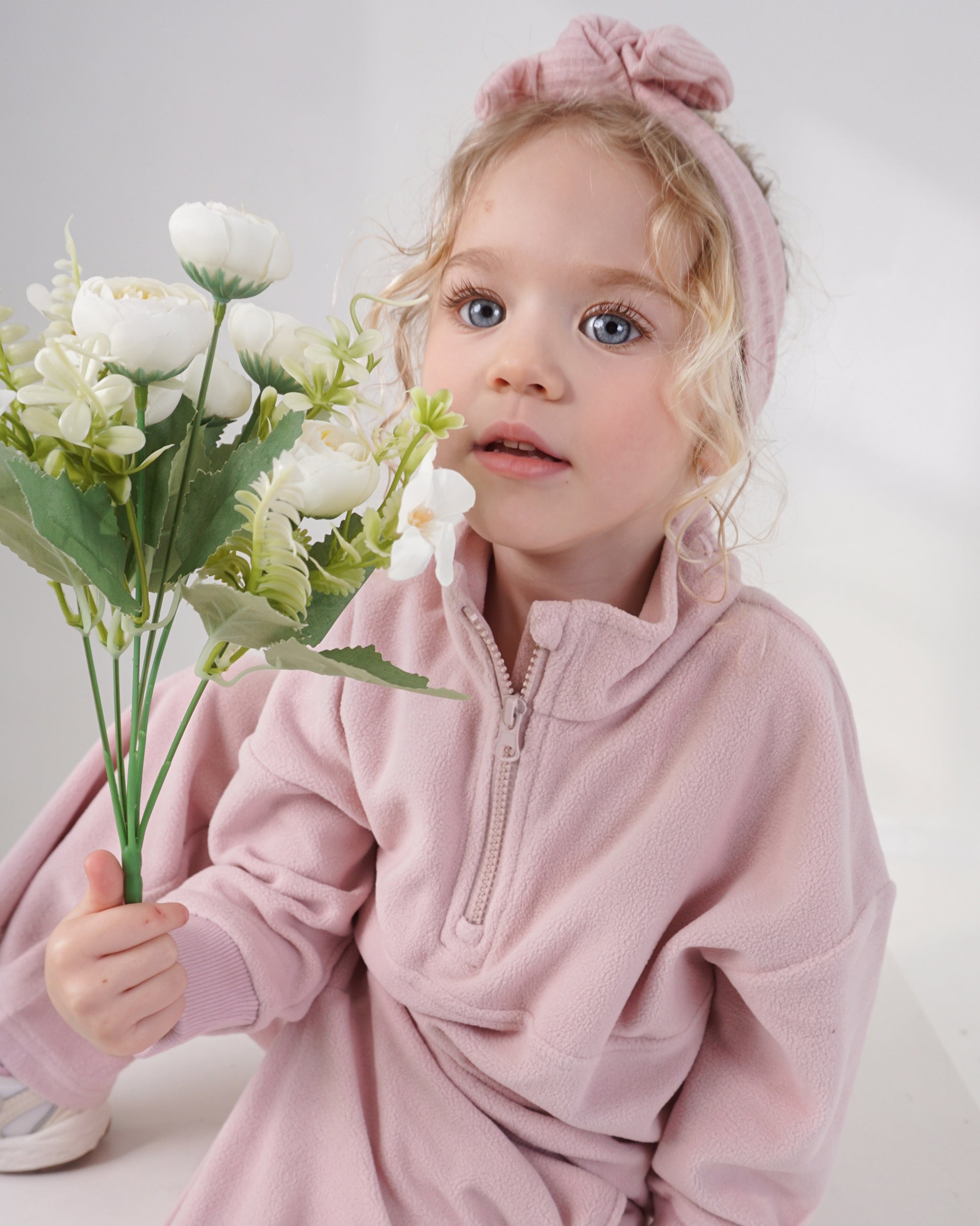 A young girl with blonde curly hair wearing a dusty pink quarter-zip fleece hoodie and a matching pink bow headband, holding a bouquet of white flowers against a light gray studio background.