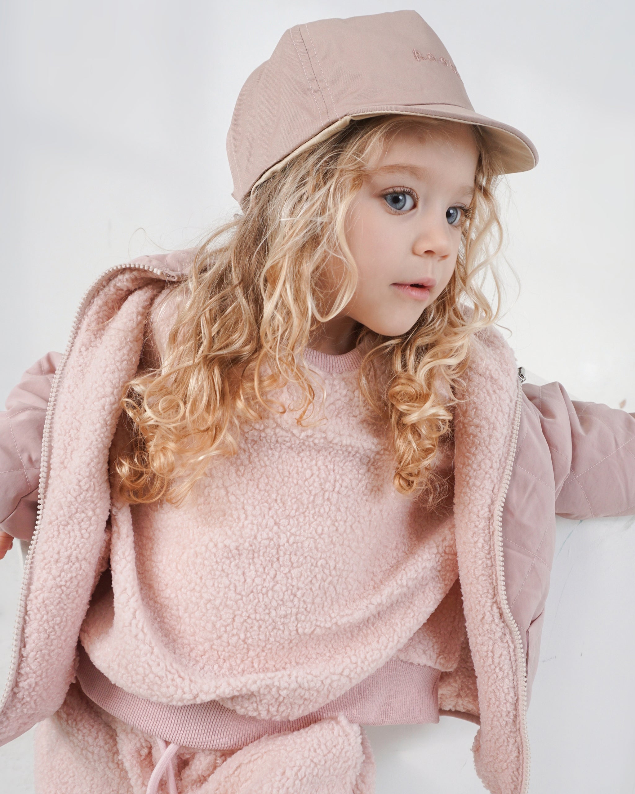 Toddler wearing a reversible cream and pink baseball cap with a pink teddy jumper and matching dusty pink reversible 4-in-1 coat and pink teddy cargo pants with a white studio backdrop.