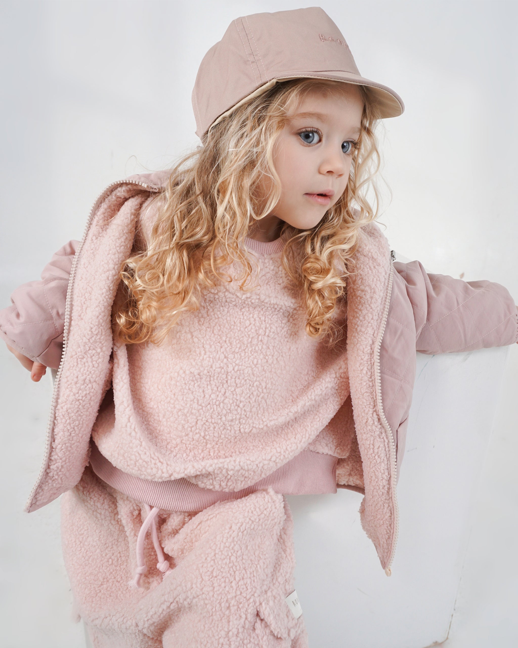 Toddler wearing a reversible cream and pink baseball cap with a pink teddy jumper and matching dusty pink reversible 4-in-1 coat and pink teddy cargo pants with a white studio backdrop.