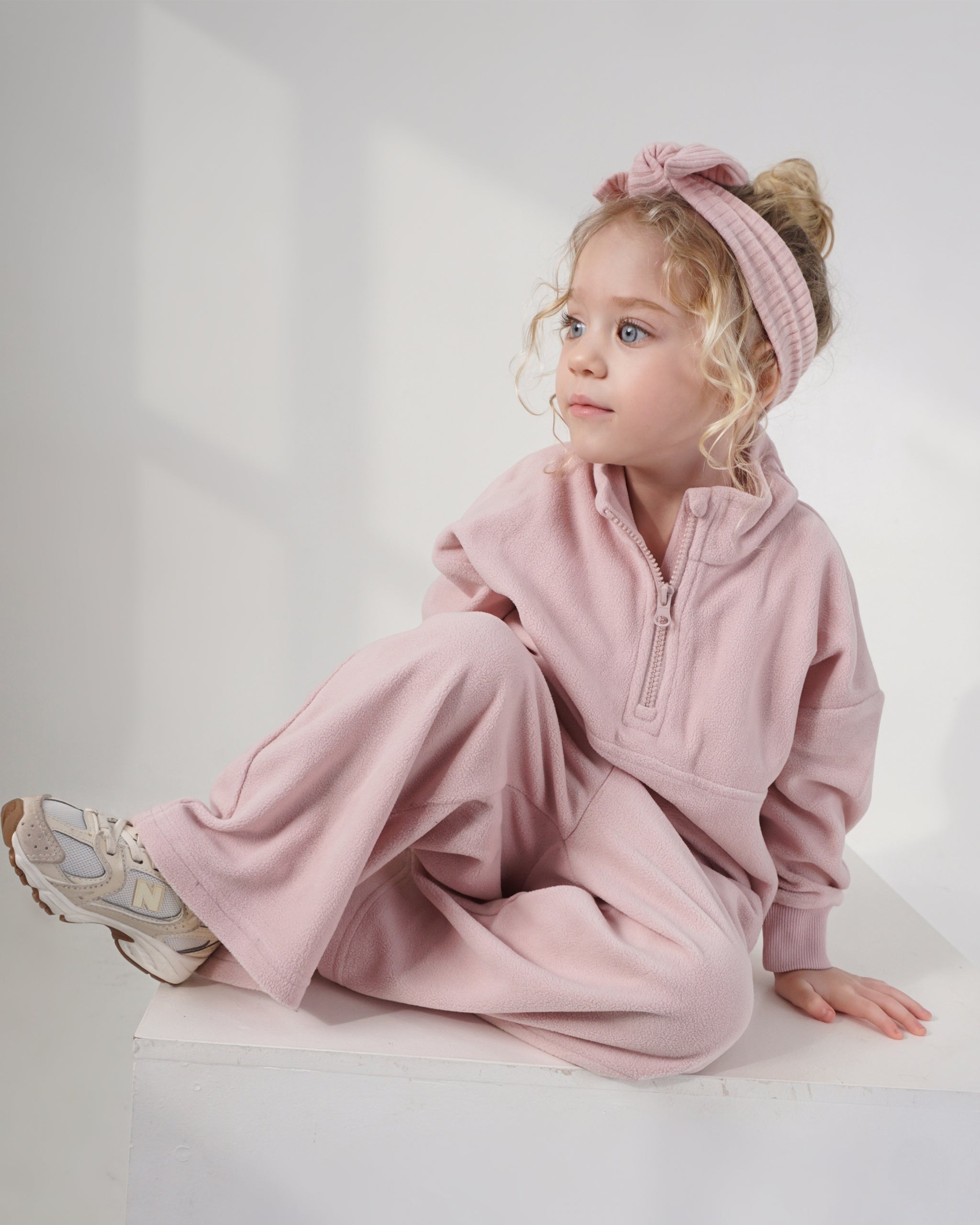 A young girl with blonde curly hair sits on a white platform wearing a dusty pink two-piece lounge set consisting of a quarter-zip long-sleeve top jumper and matching wide-leg pants, along with a pink ribbed bow headband and sneakers.