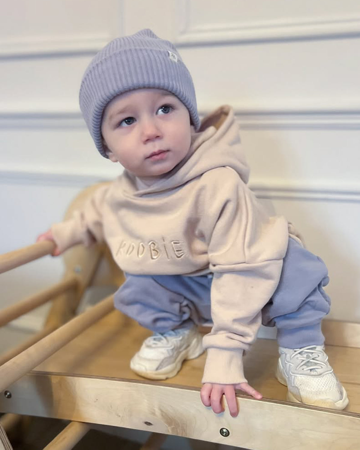 Child wearing a beige hoodie with text, light blue pants, and a matching beanie on a wooden chair.