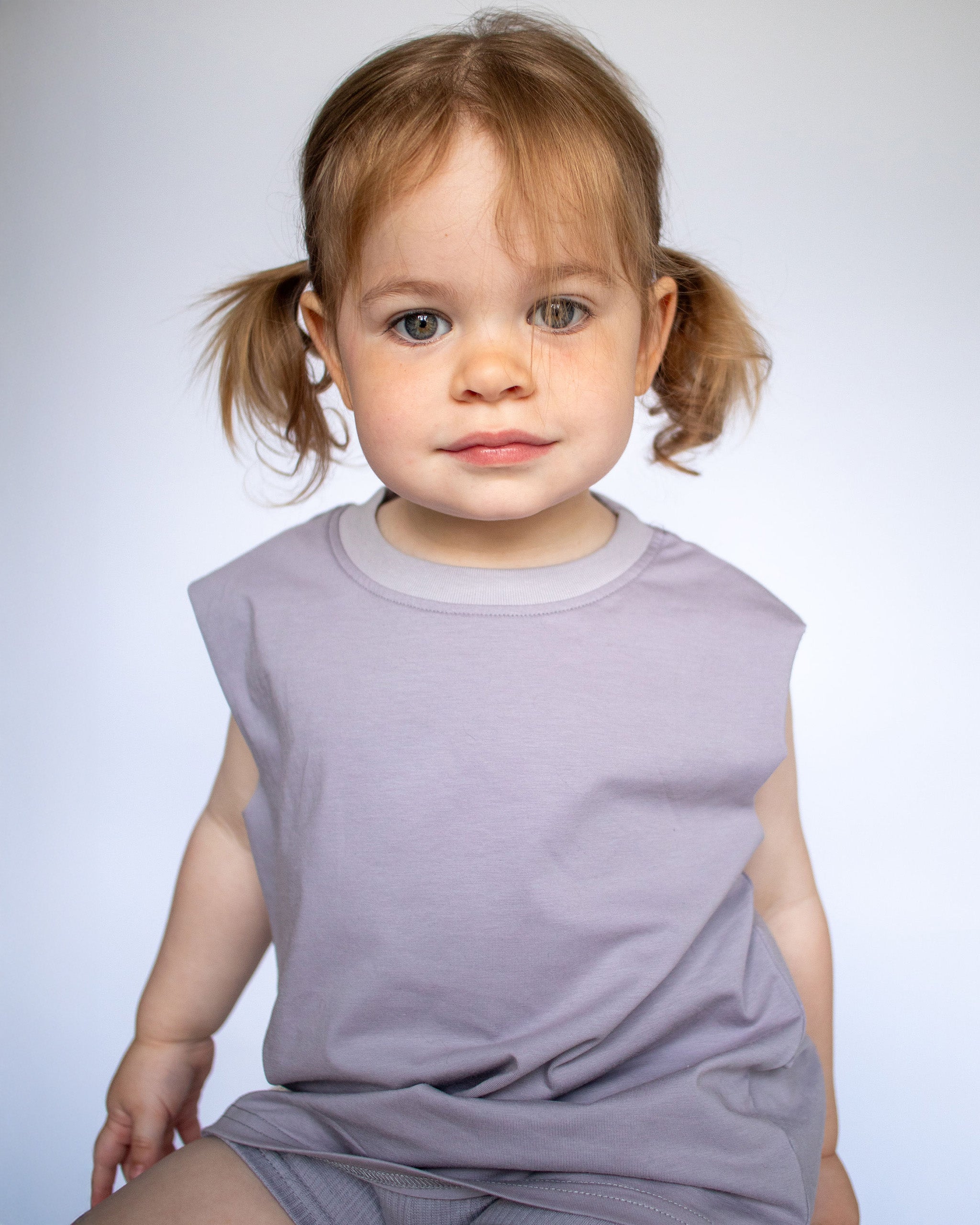 A toddler girl with light brown hair in pigtails wearing a sleeveless light blue-gray Singlet t-shirt and matching Ribbed Cycling Shorts, sitting against a light neutral gray studio background.