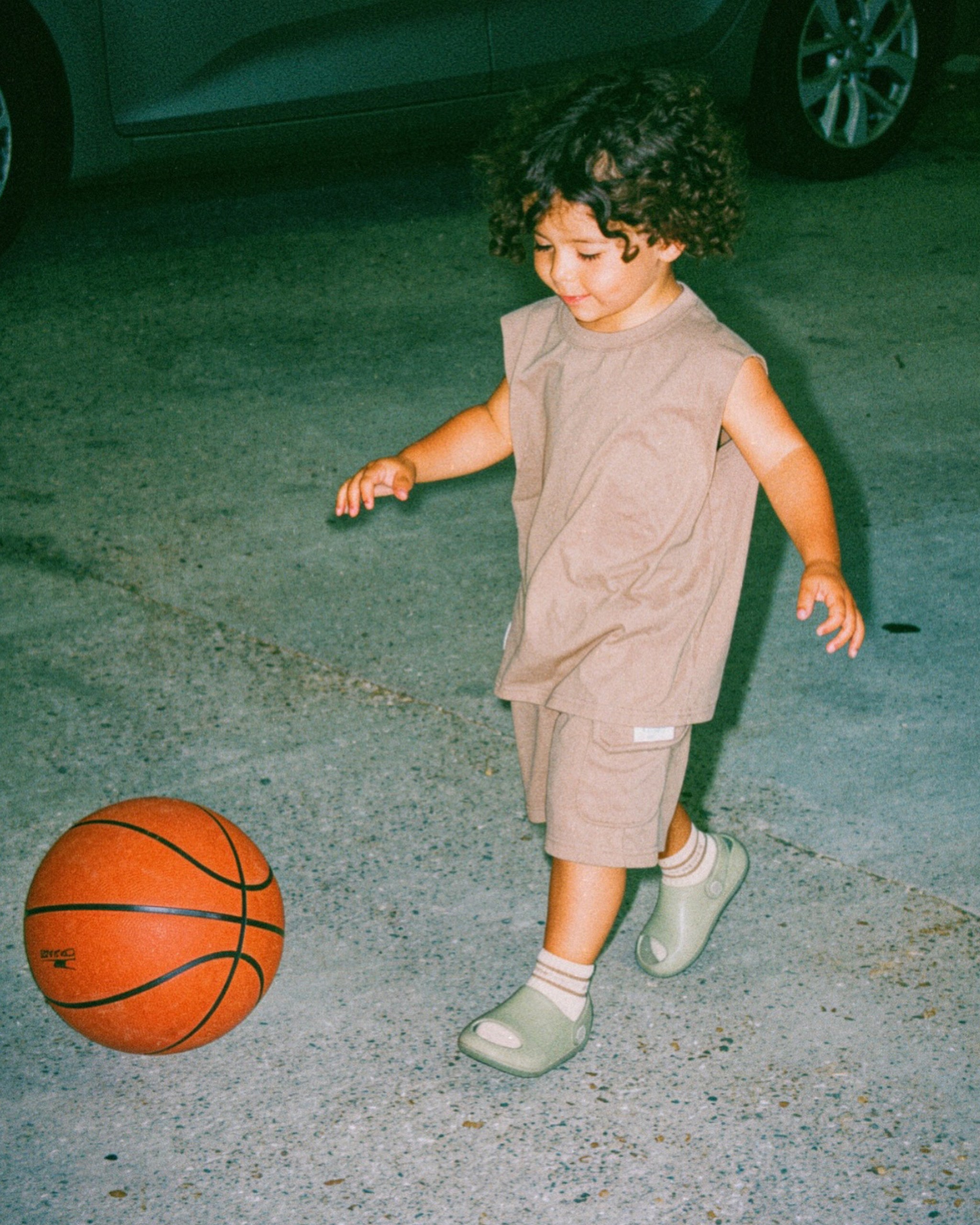 A toddler with curly dark hair wearing a matching beige (CHOCCY BiCCY) Singlet sleeveless top and Cargo Shorts outfit, walking outdoors on a concrete surface next to an orange basketball.