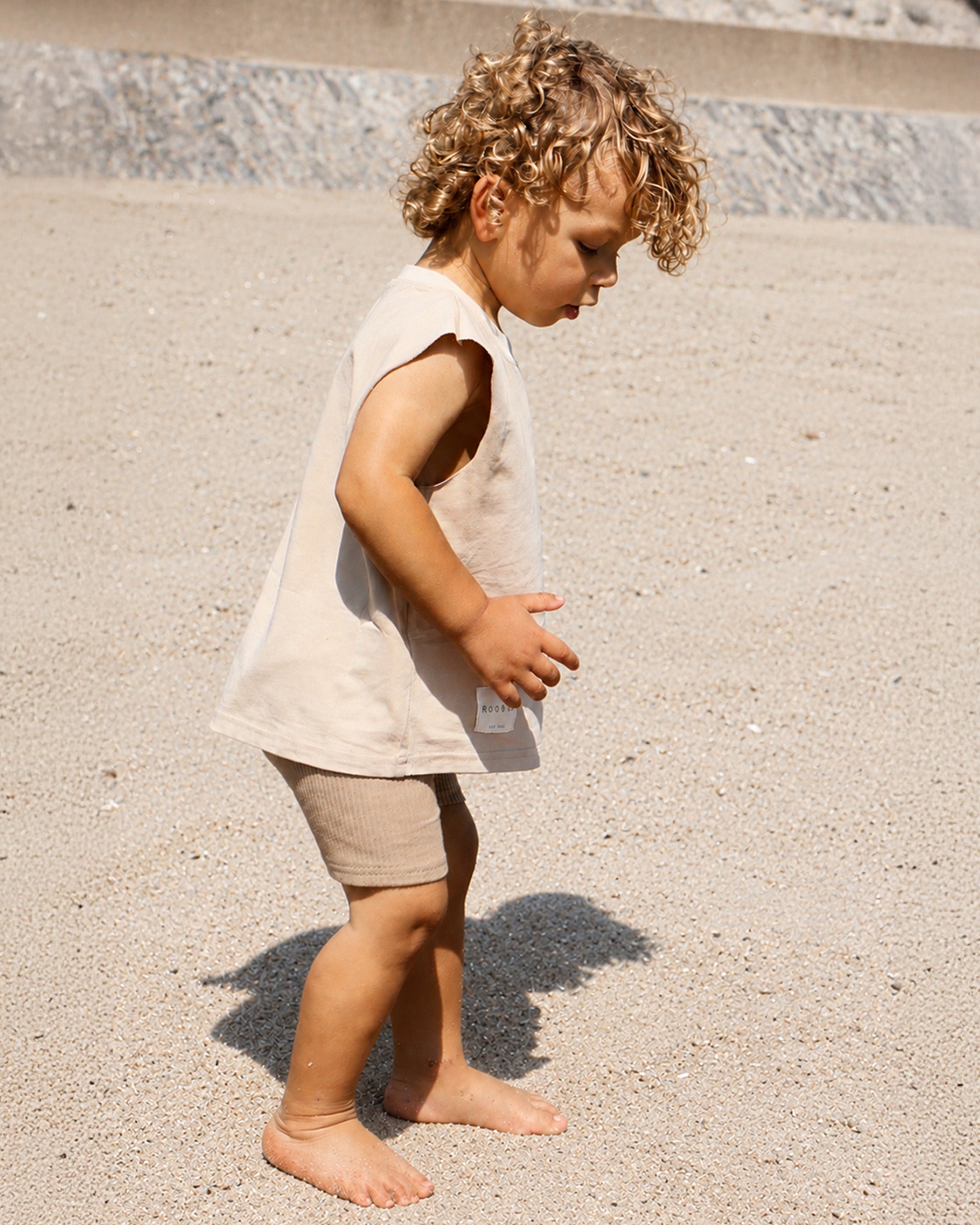 A toddler with curly blond hair stands barefoot on a sandy surface, wearing a sleeveless light beige Singlet top and tan Ribbed Cycling Shorts.