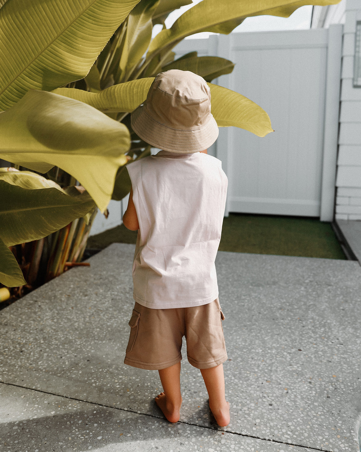 A toddler seen from behind wearing a light beige Reversible Bucket Hat, an oversized cream sleeveless Singlet top, and tan Cargo Shorts, standing barefoot on a concrete patio in a backyard setting.