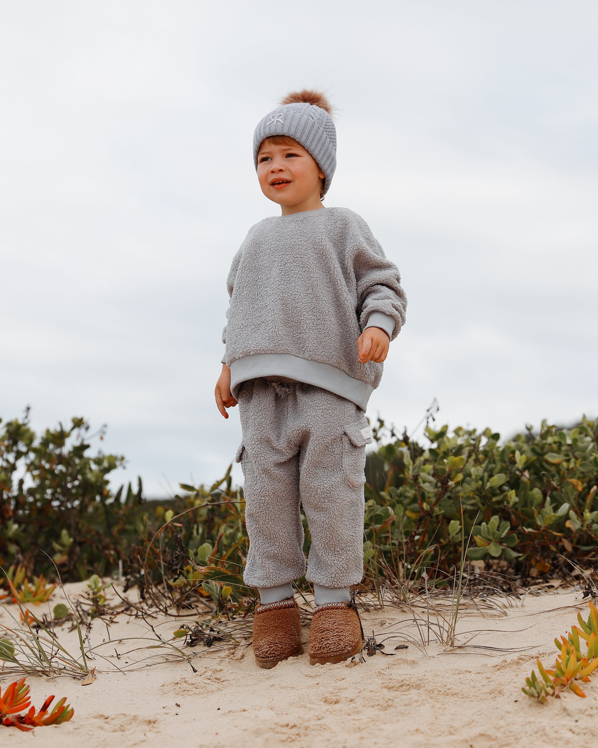 A toddler stands on sandy ground outdoors wearing a light gray-blue knit bobble beanie with a pom-pom, a matching neutral blue long-sleeve fleece teddy jumper and blue-gray teddy jogger-style cargo pants, and brown ugg boots. Low green shrubs and a pale cloudy sky are in the background.