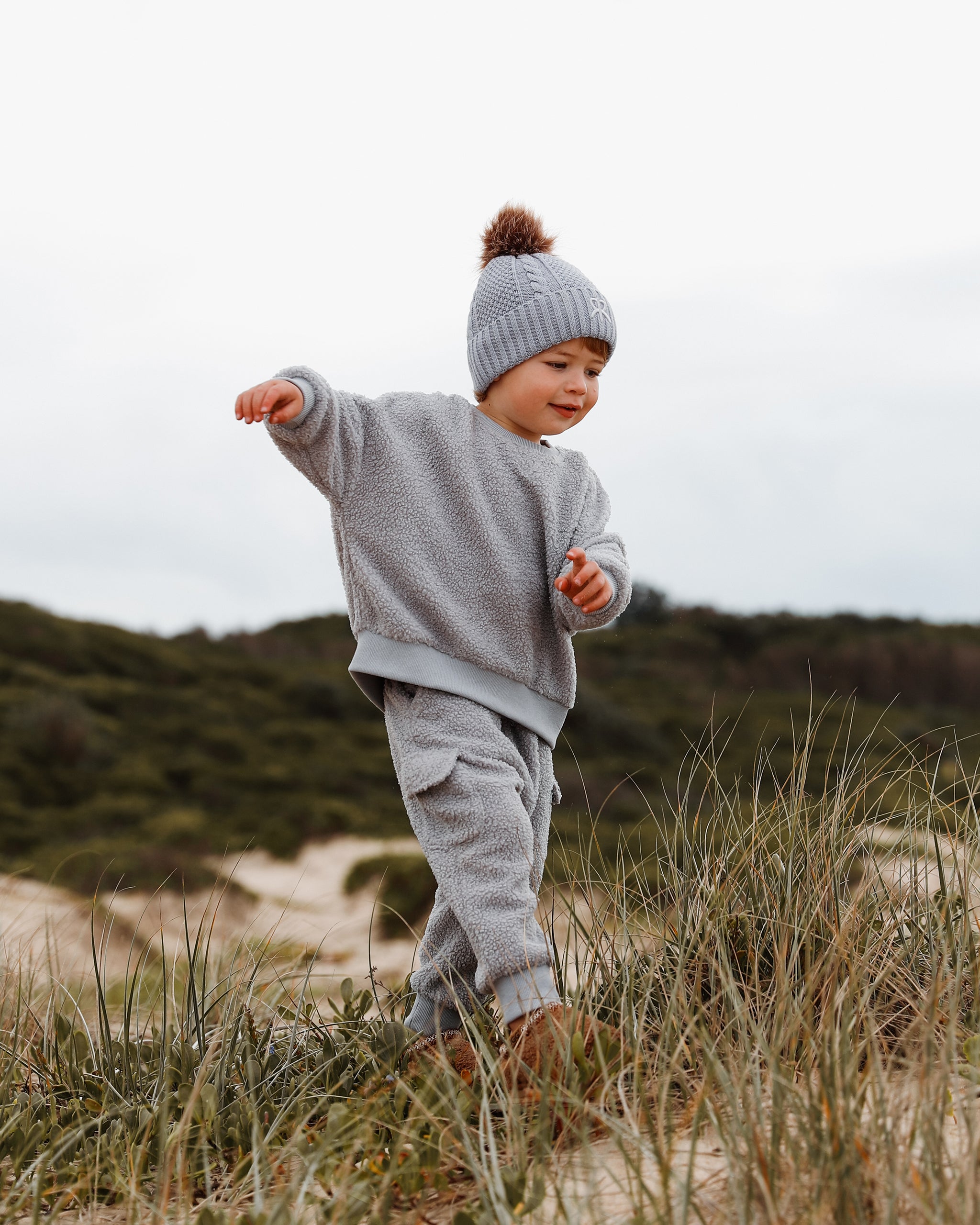 A toddler walking outdoors on sandy dunes wearing a light gray-blue teddy jumper, matching cargo jogger pants, and a blue-gray knit bobble beanie with a brown pom-pom.