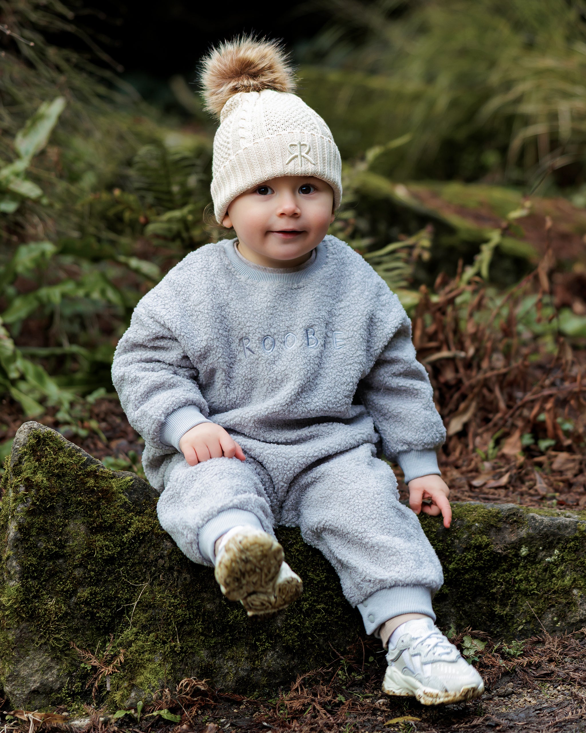 Child wearing a light blue fleece onesie style one-piece with a cream knit bobble beanie hat, sitting on a log in a forest setting.