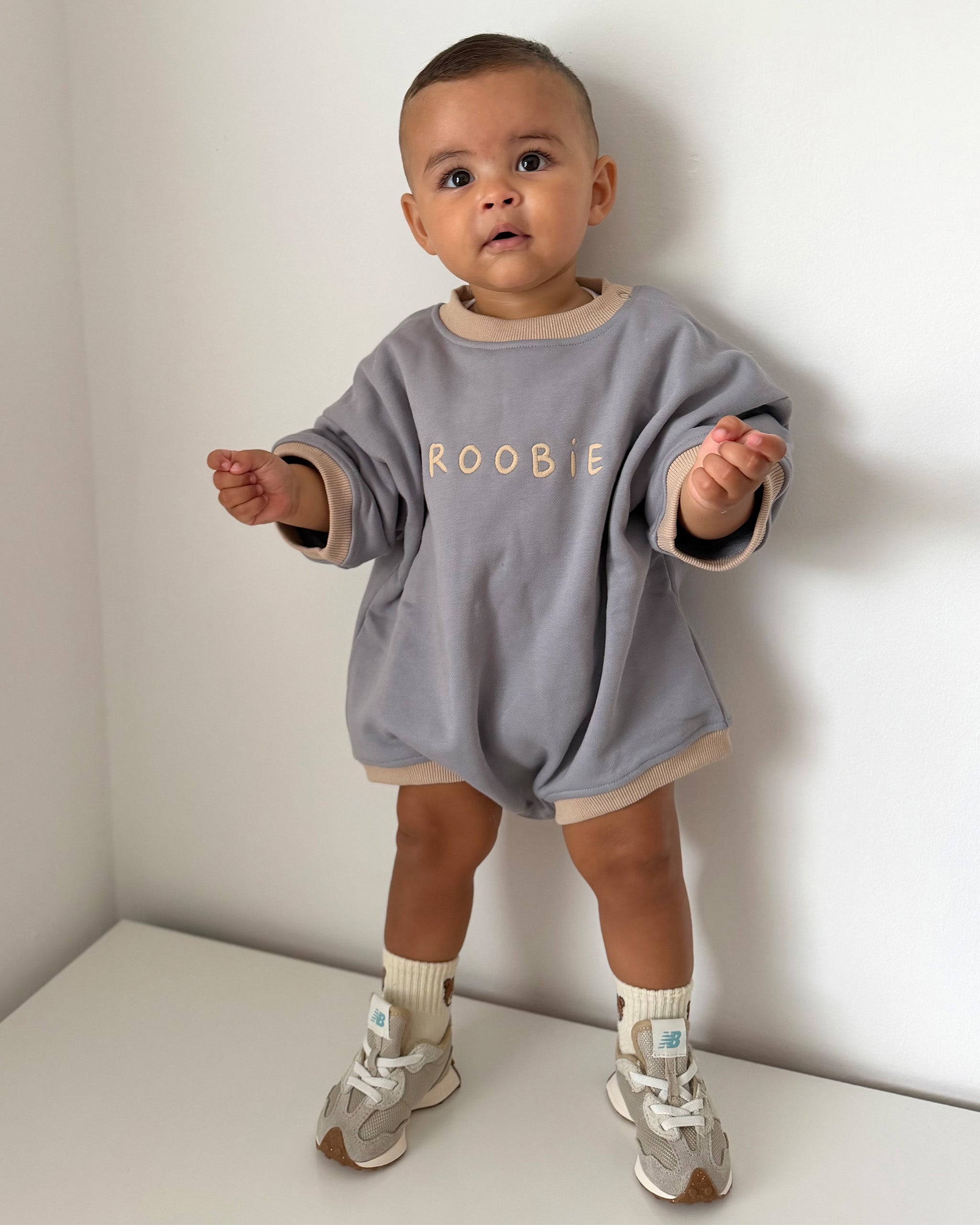 A toddler stands in a white room corner wearing a neutral gray-blue Oversized Romper with beige trim and the word 'ROOBIE' printed across the chest in beige. The child is also wearing beige socks and gray sneakers.