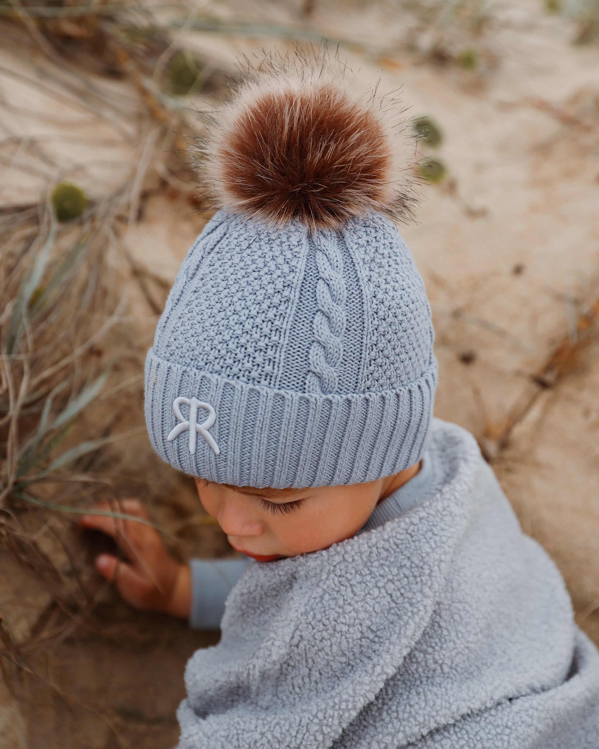 A baby wearing a light blue cable-knit bobble beanie with a brown faux fur pom-pom and embroidered logo detail, wearing in a matching teddy fleece jumper in a sandy outdoor setting.