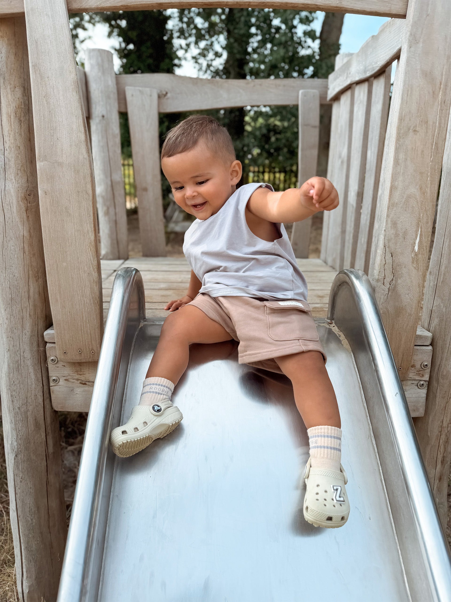 A toddler sitting on a metal slide in an outdoor playground, wearing a blue (BONDi BREEZE) sleeveless Singlet top, beige (CHOCCY BiCCY) Cargo Shorts, cream and blue striped ROOBiE crew socks, and cream-colored Croc-style shoes.