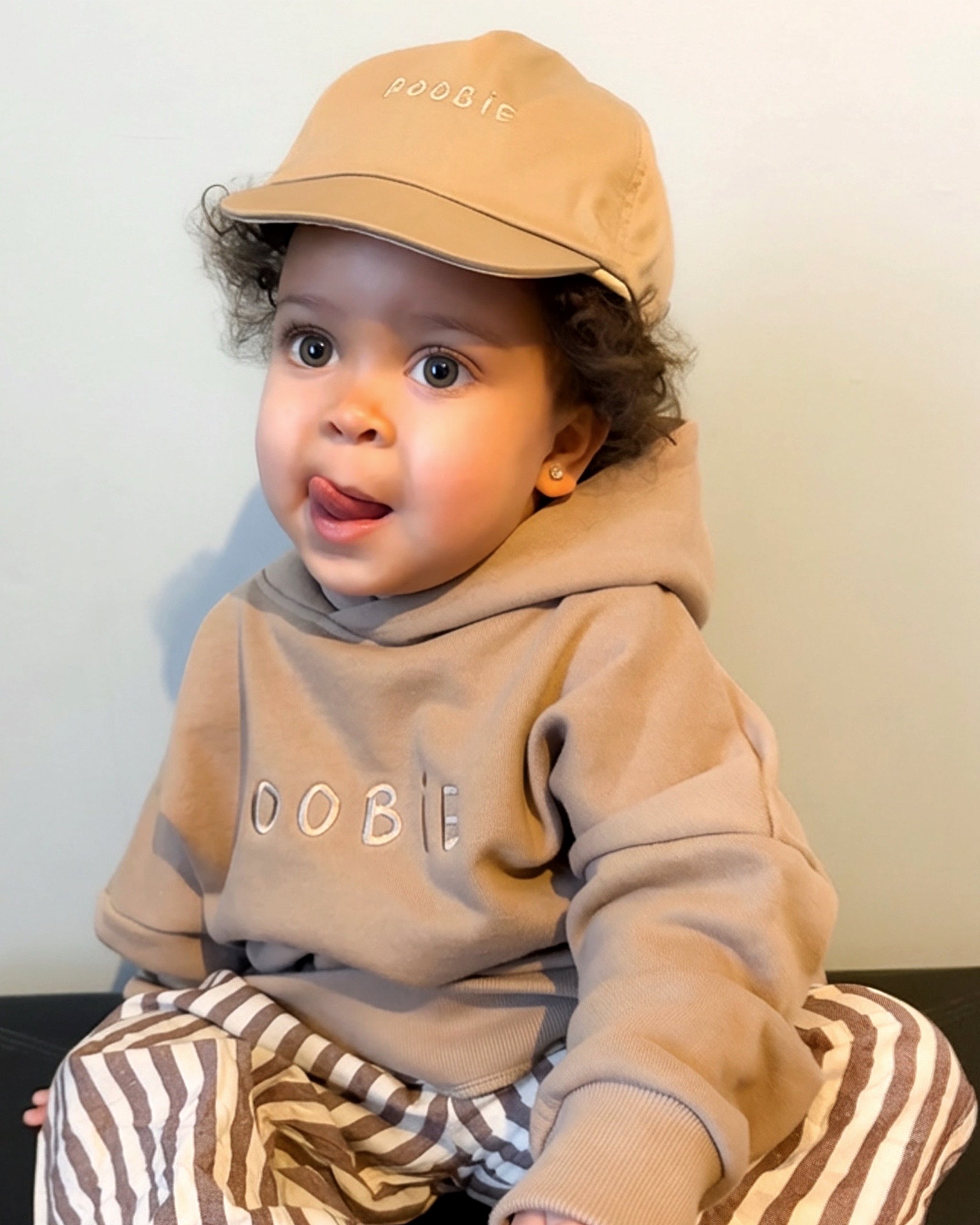 Child wearing a beige reversible cap and oversized hoodie with 'ROOBiE' branding, sitting on a striped cushion.