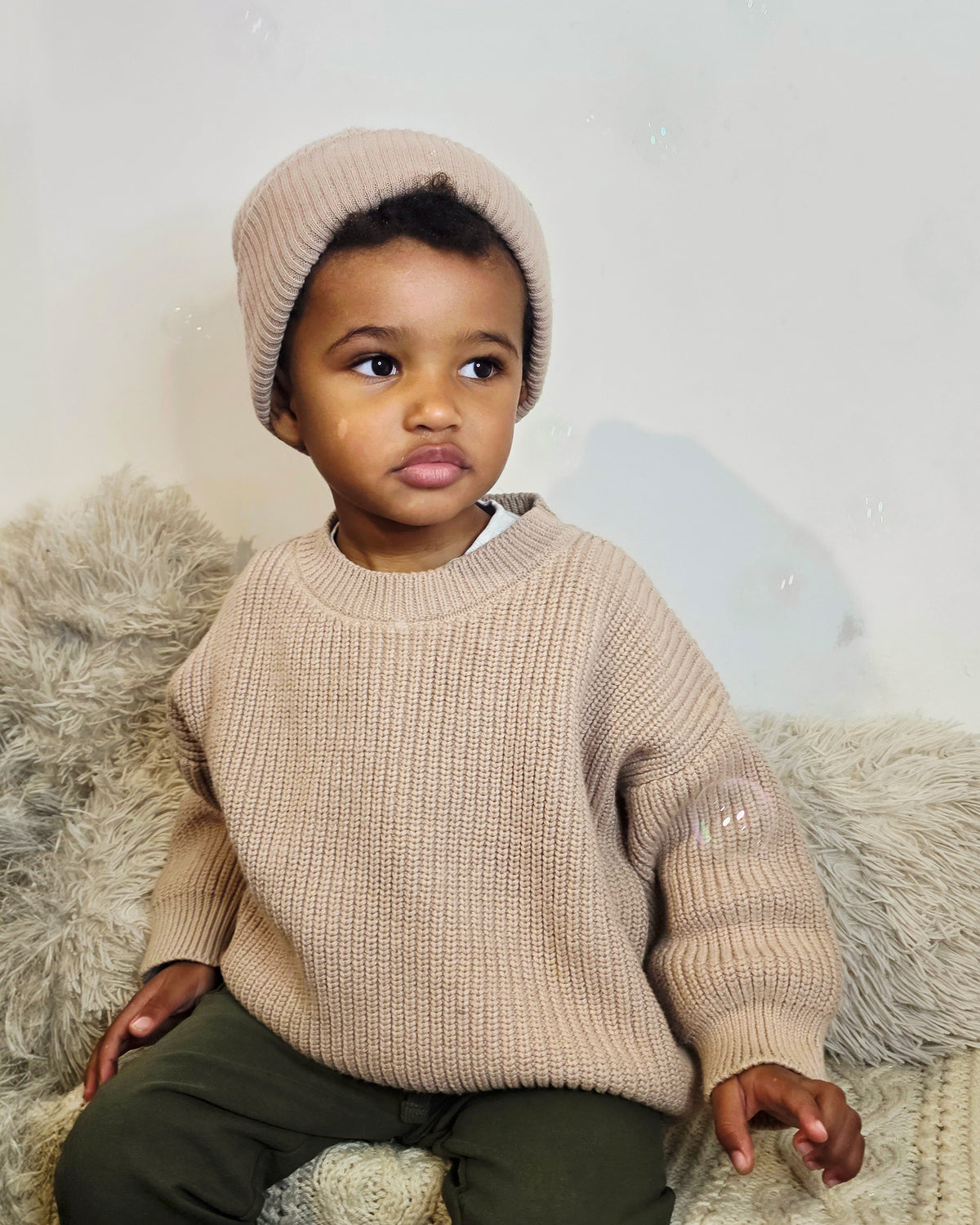 Child wearing a brown chunky knit sweater and ribbed cotton beanie sitting on a fluffy surface.